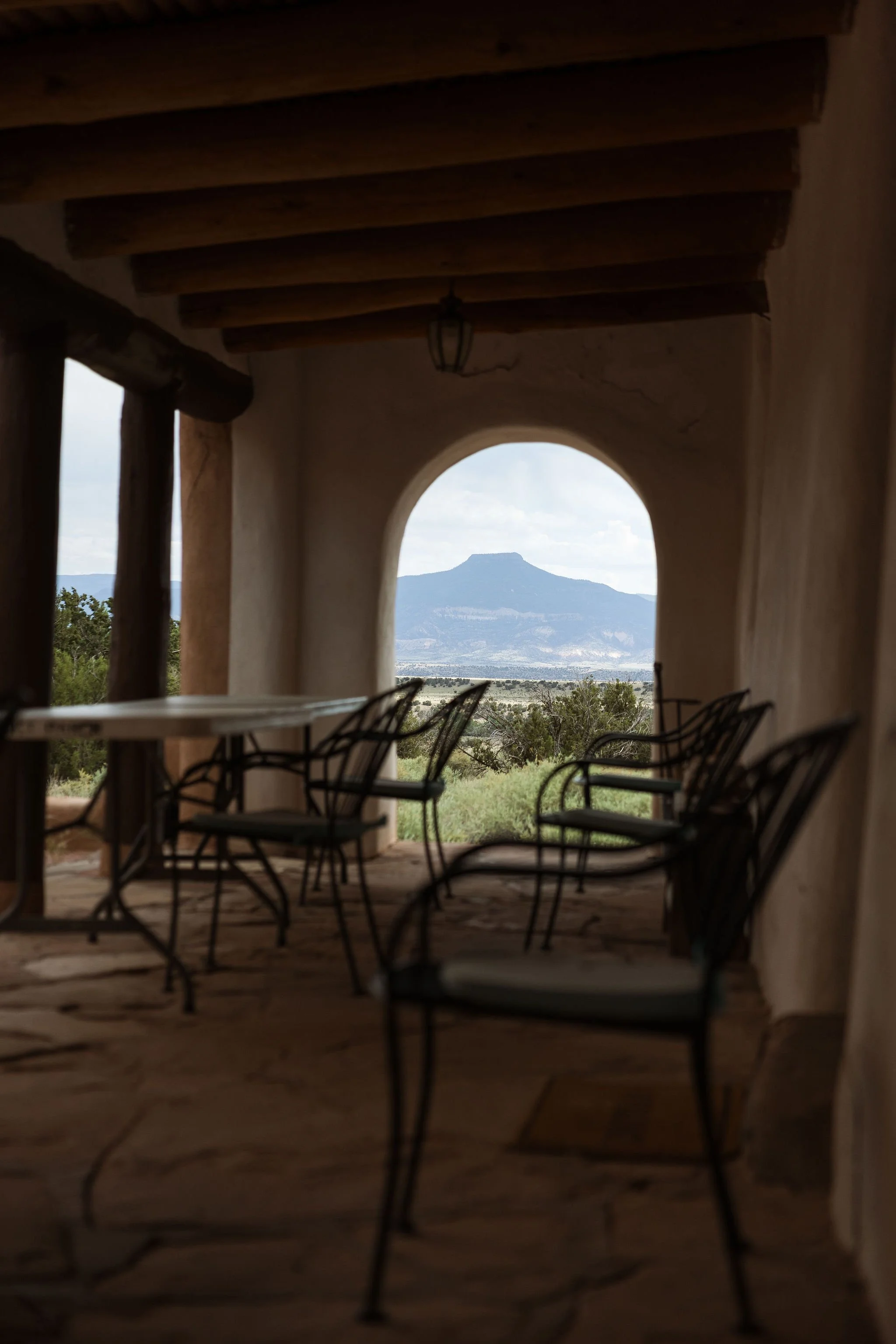 Looking through an arched window with wrought iron chairs inside a rustic room, a distant mountain is visible under a cloudy sky.