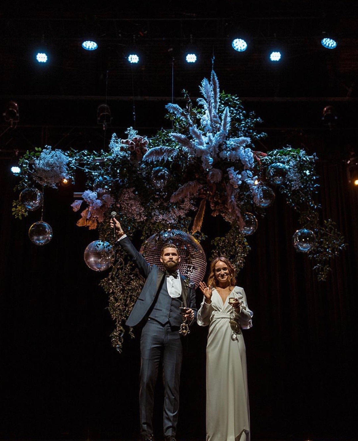 A bride and groom standing under a large floral arrangement with hanging disco balls, on a stage with blue lights.