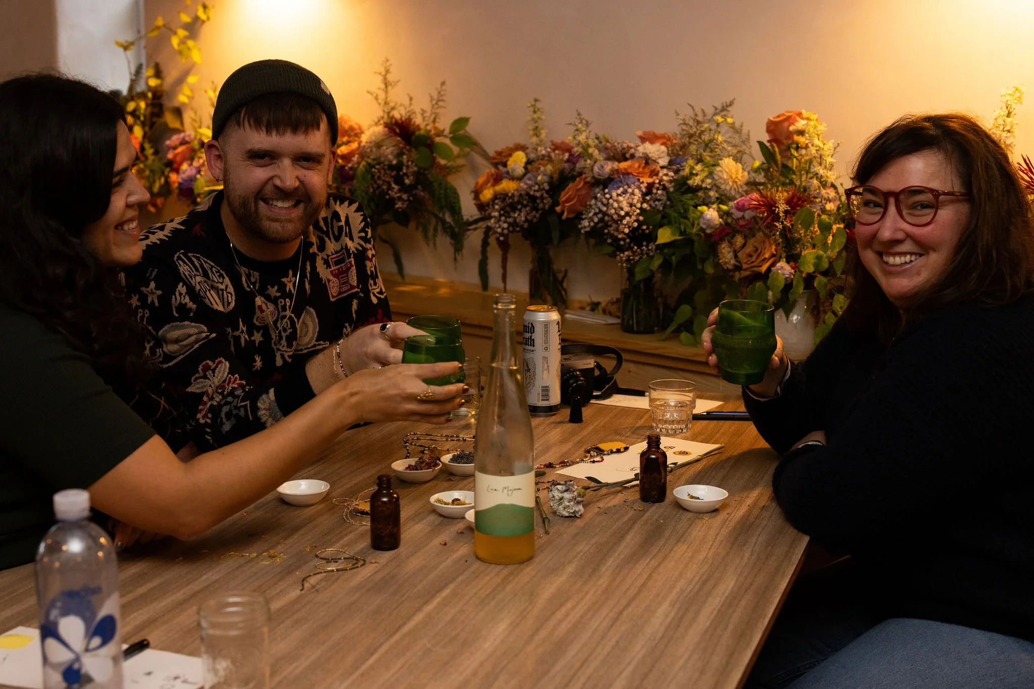 Three friends sitting at a wooden table, smiling and clinking glasses with green drinks, surrounded by flowers and art supplies.