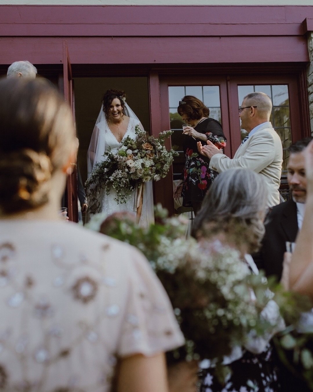 A bride standing at her wedding reception outside, smiling and holding a large bouquet of flowers, surrounded by guests who are clapping and celebrating.
