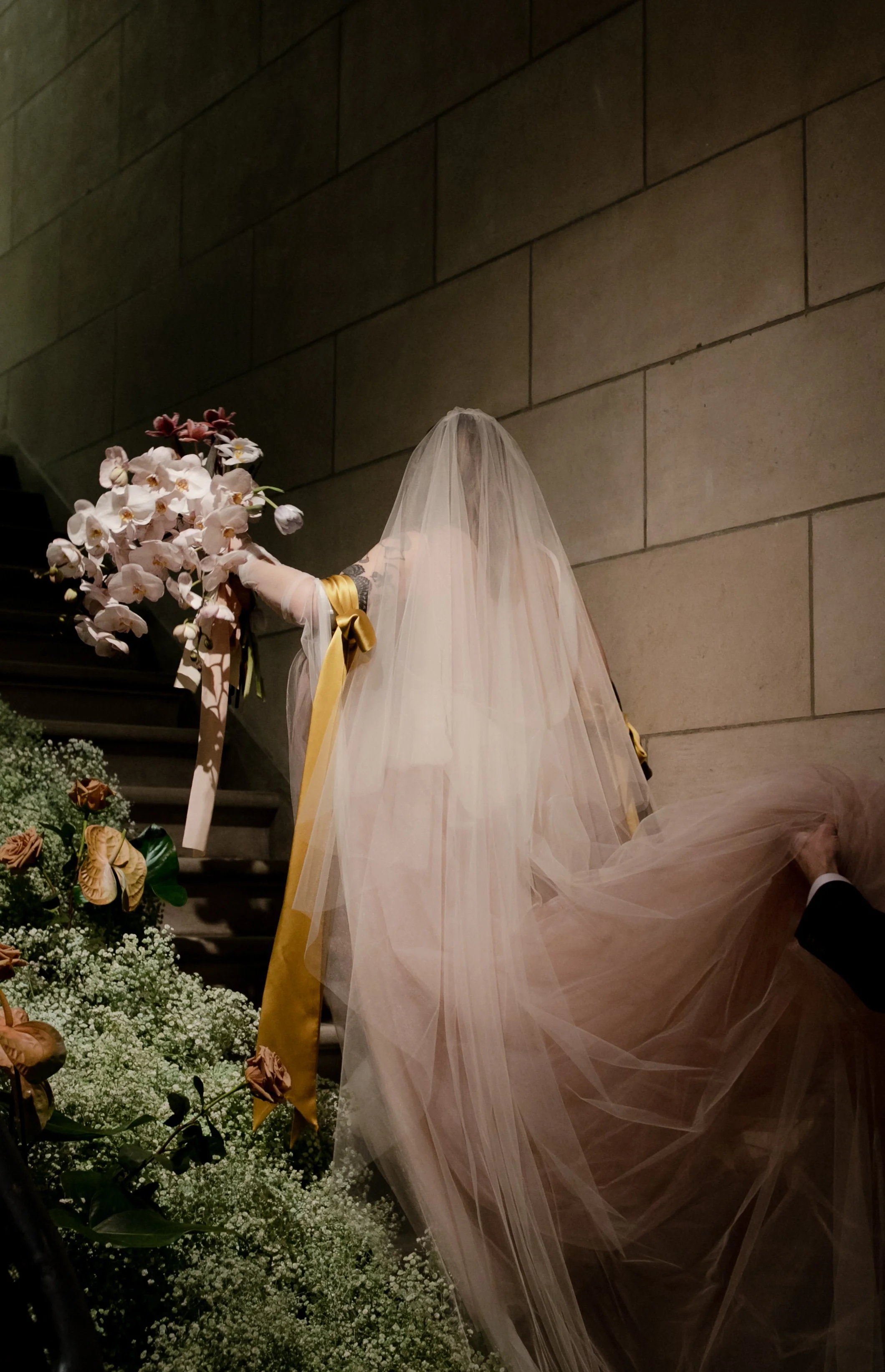 A bride in a wedding dress with a long veil sits on steps, holding a bouquet of pink orchids, surrounded by flowers.