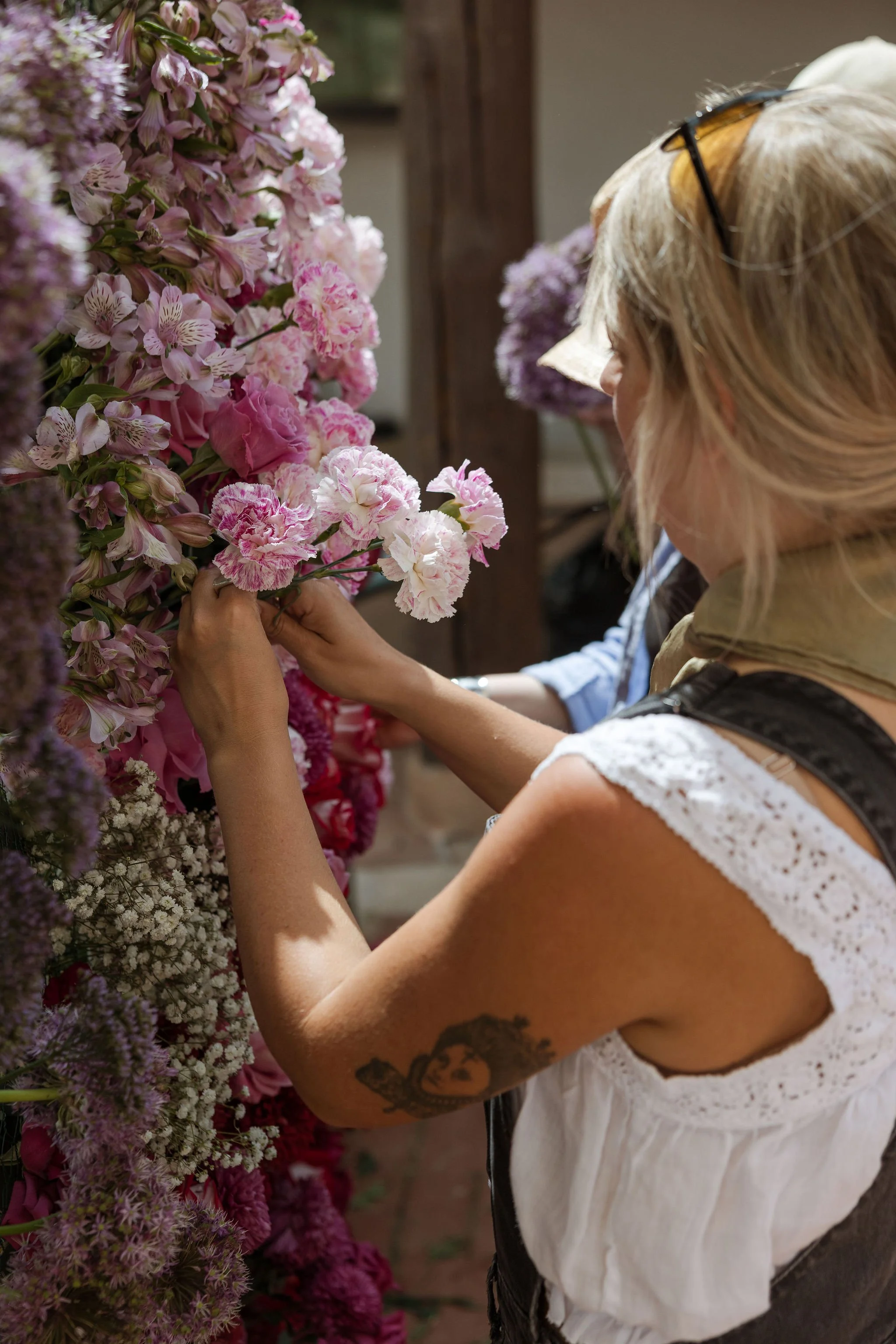 A woman with a tattoo on her arm arranging pink and purple flowers on a floral wall in a workshop.