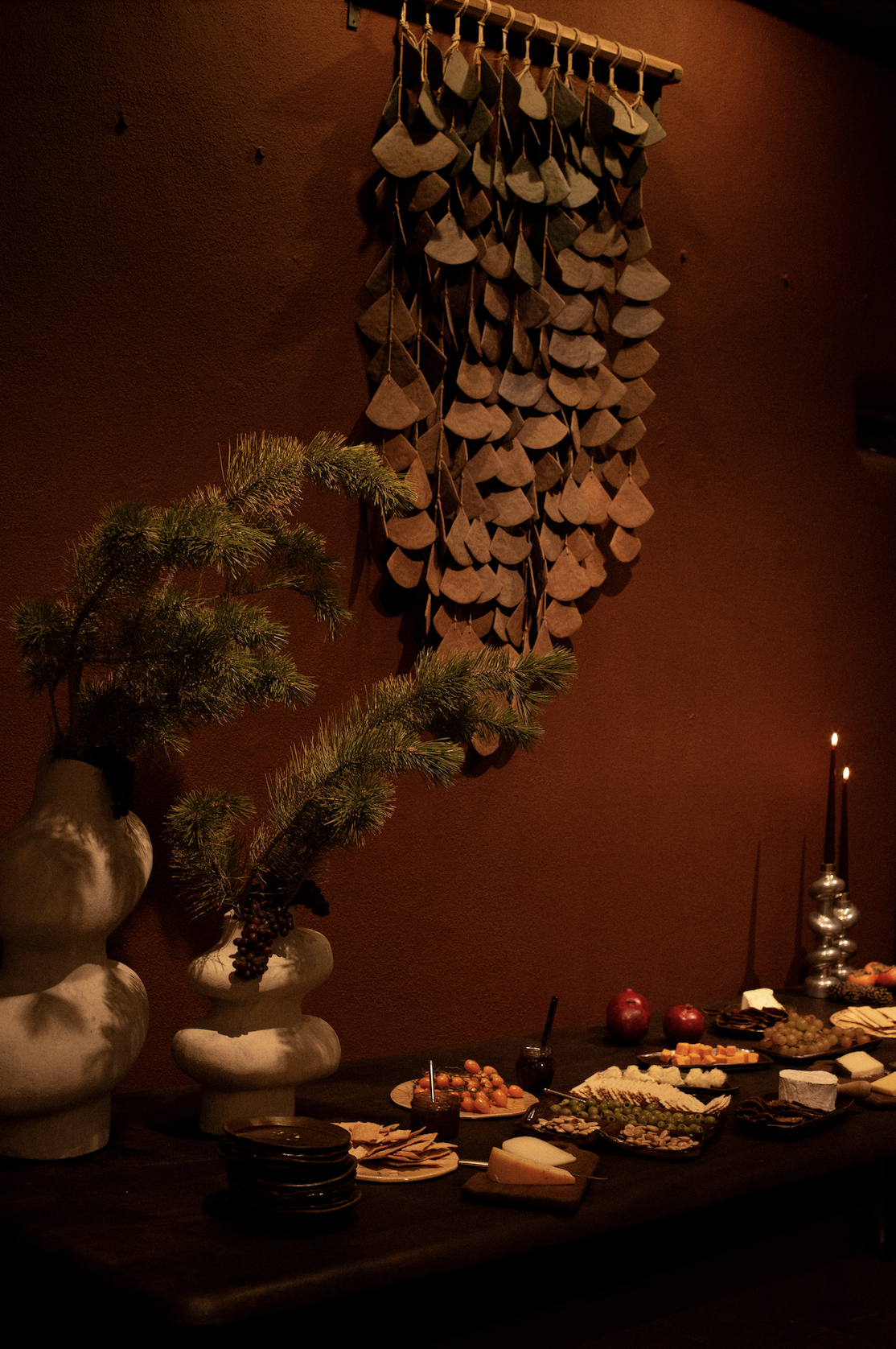 A table set with cheeses, grapes, crackers, and small glasses of drinks, flanked by vases with pine branches, against a warm, burgundy-colored wall decorated with hanging leather or wooden ornaments.