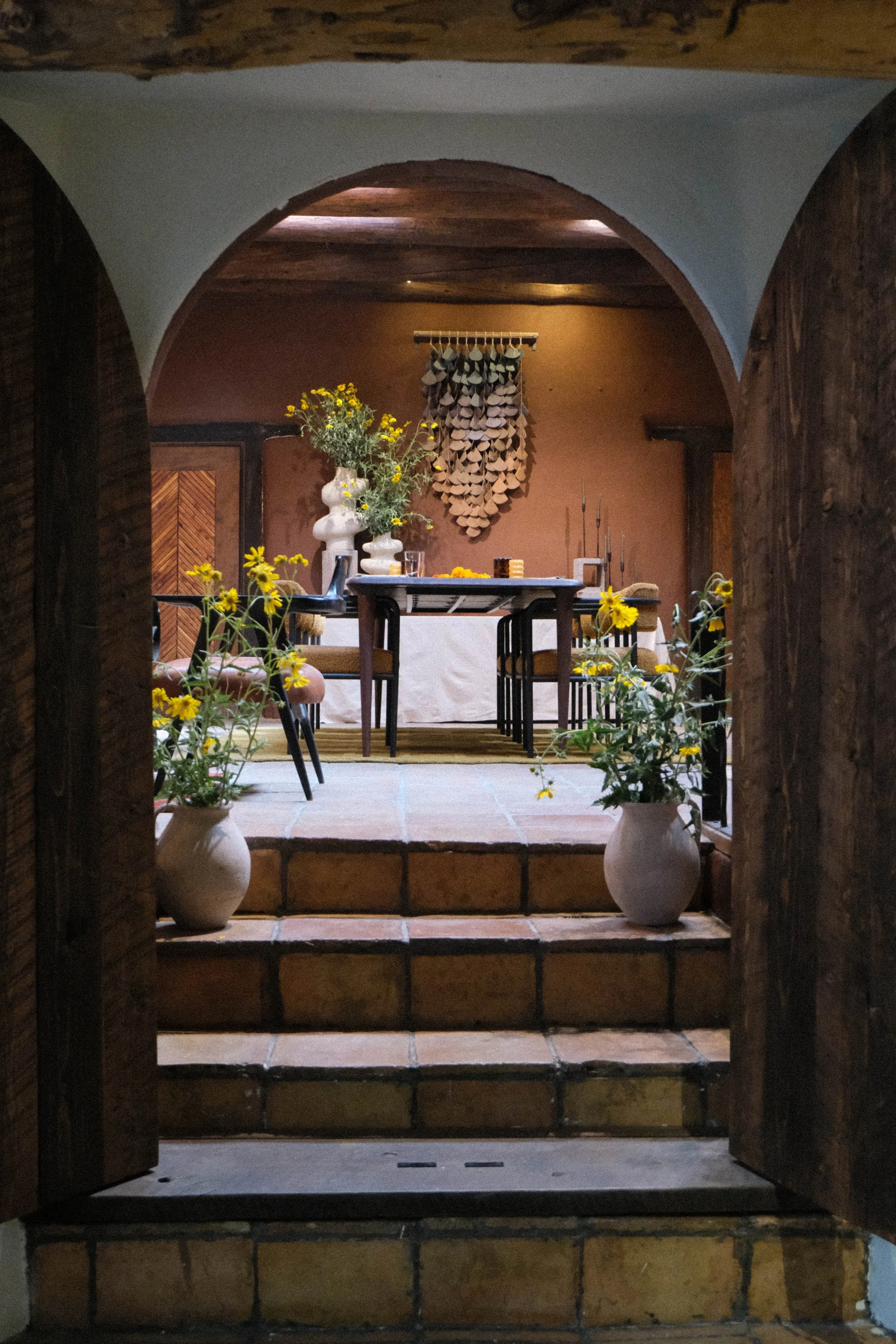 A cozy indoor space with rustic decor, featuring a dining table with chairs, potted yellow flowers, wall art, and warm lighting.