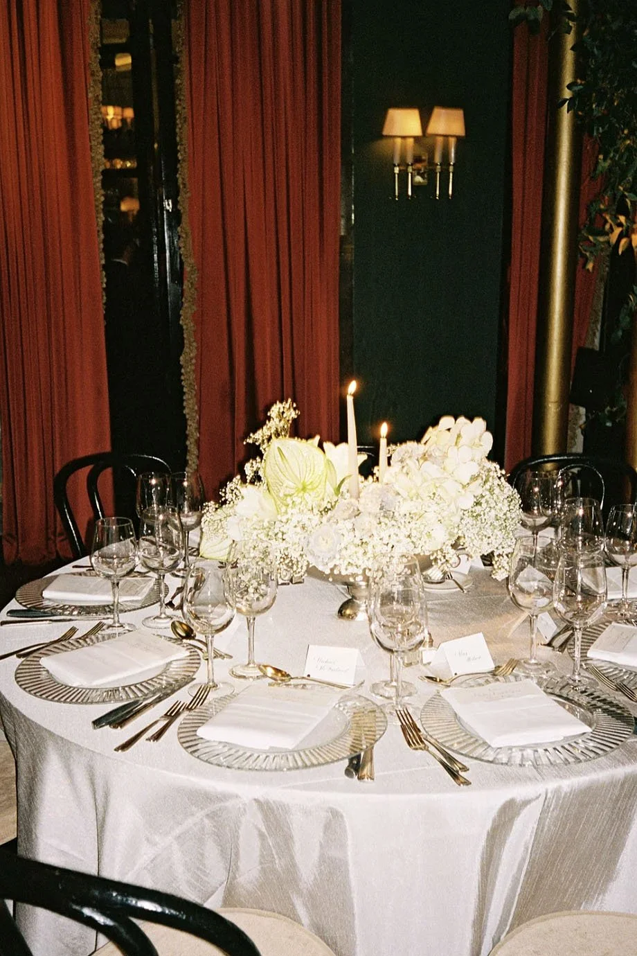 Round table set for a formal dinner with white tablecloth, elegant glassware, gold flatware, white napkins, and a floral centerpiece with white flowers and two lit candles, in a room with dark walls, red curtains, and wall-mounted lamps.