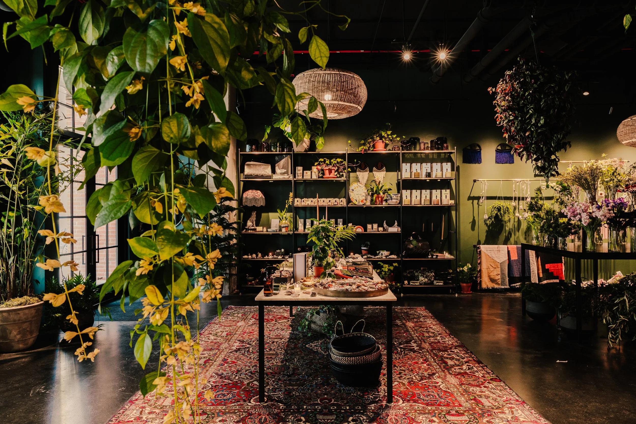 Interior of a boutique shop with green walls, multiple plants, a central table with assorted items, Shelf filled with various products, and floral arrangements, illuminated by hanging woven lamps.