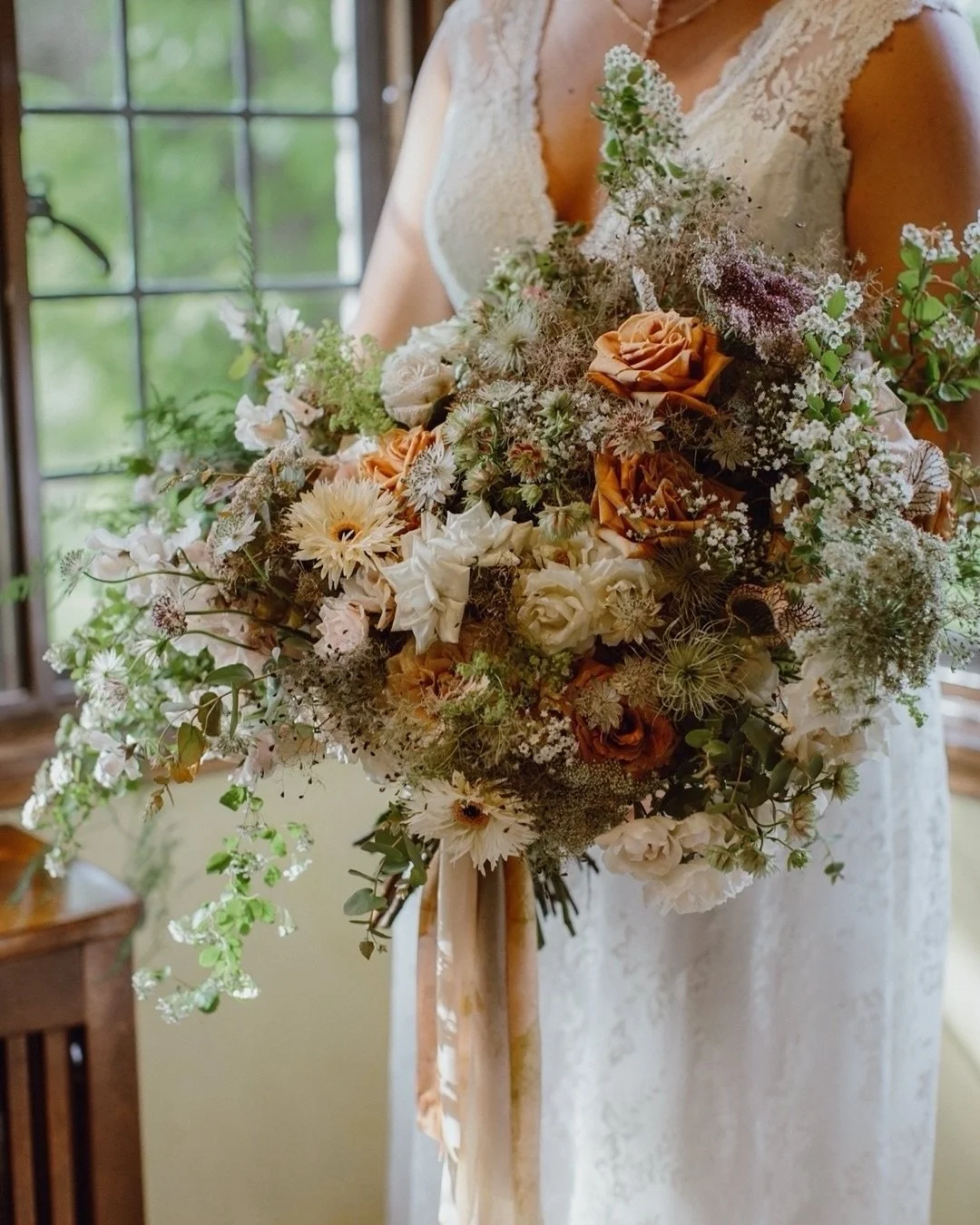 A woman in a lace wedding dress holding a large bouquet of flowers with peach, white, and green blooms, tied with a peach satin ribbon.