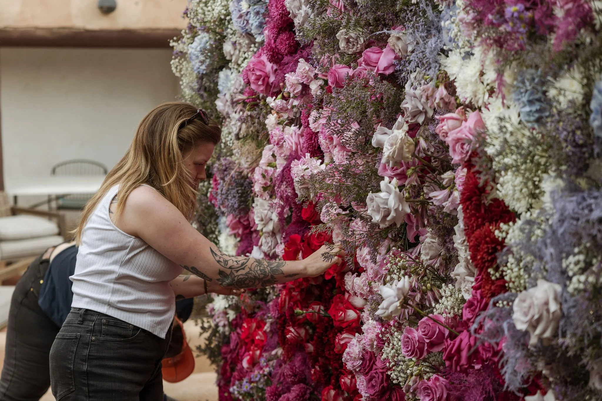 Woman with tattoos on her arm touches a large floral wall of pink, purple, white, and red flowers.