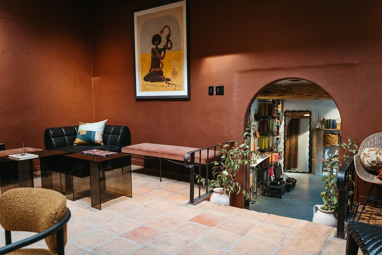 Interior of a cozy room with a reddish wall, black leather sofa with cushions, and a small side table. An art piece depicting a person with a snake hangs on the wall. Potted plants are placed near an arched opening leading to a room with shelves, boo