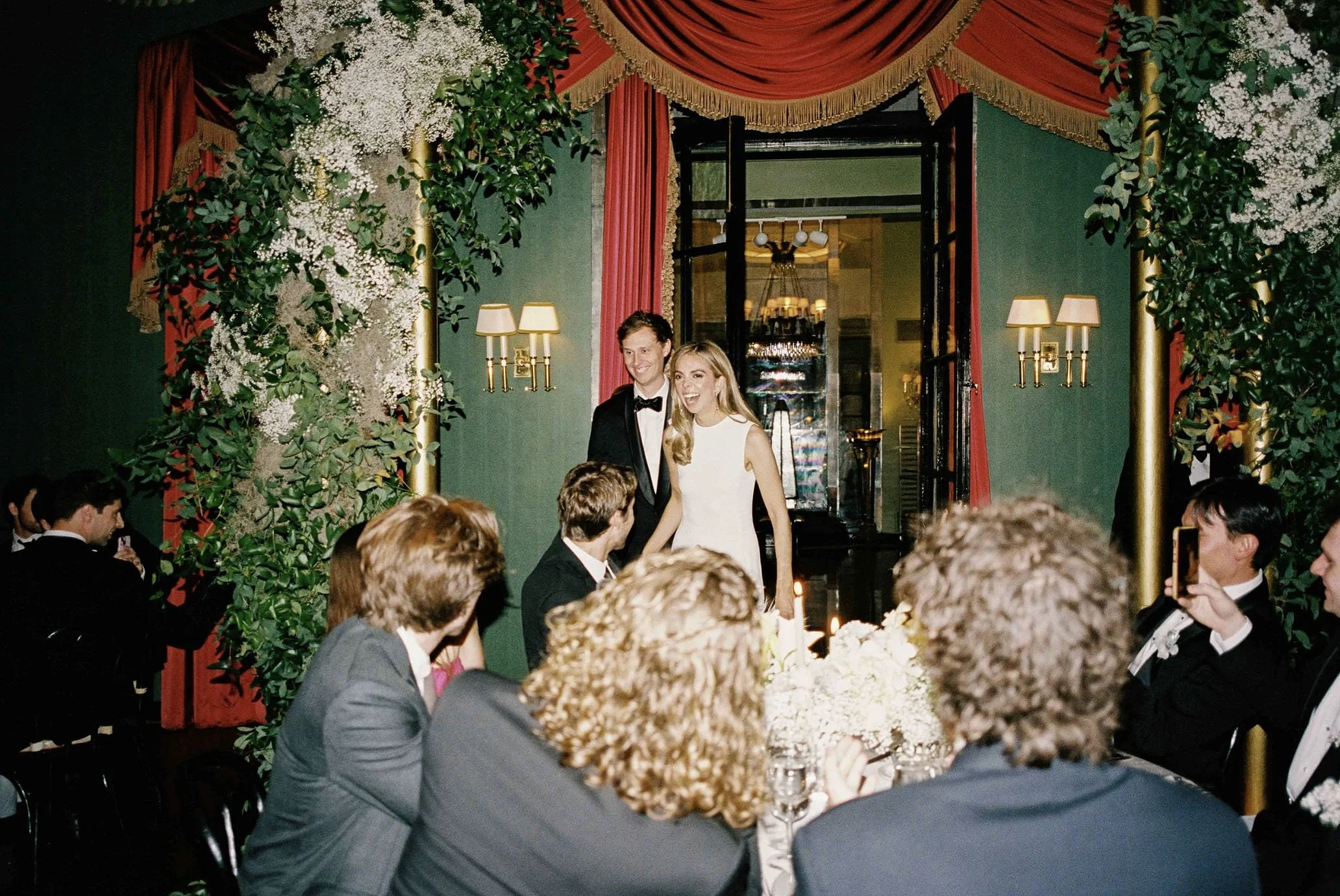 A wedding reception with a bride and groom walking into a decorated room filled with guests. The background features dark green walls, red curtains, and floral arrangements with white flowers and greenery. Guests are seated around tables, some taking