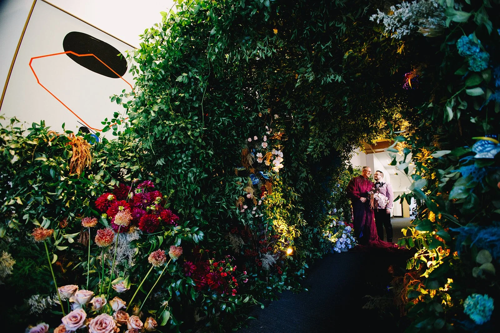 Two men standing and smiling under a floral archway decorated with various colorful flowers, greenery, and dim lighting.