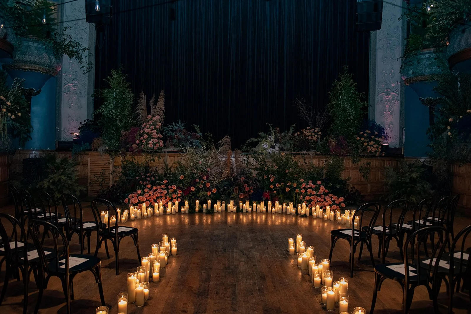 Indoor venue decorated for a wedding or event with a floral backdrop, candlelit circle on the floor, and chairs arranged on either side.