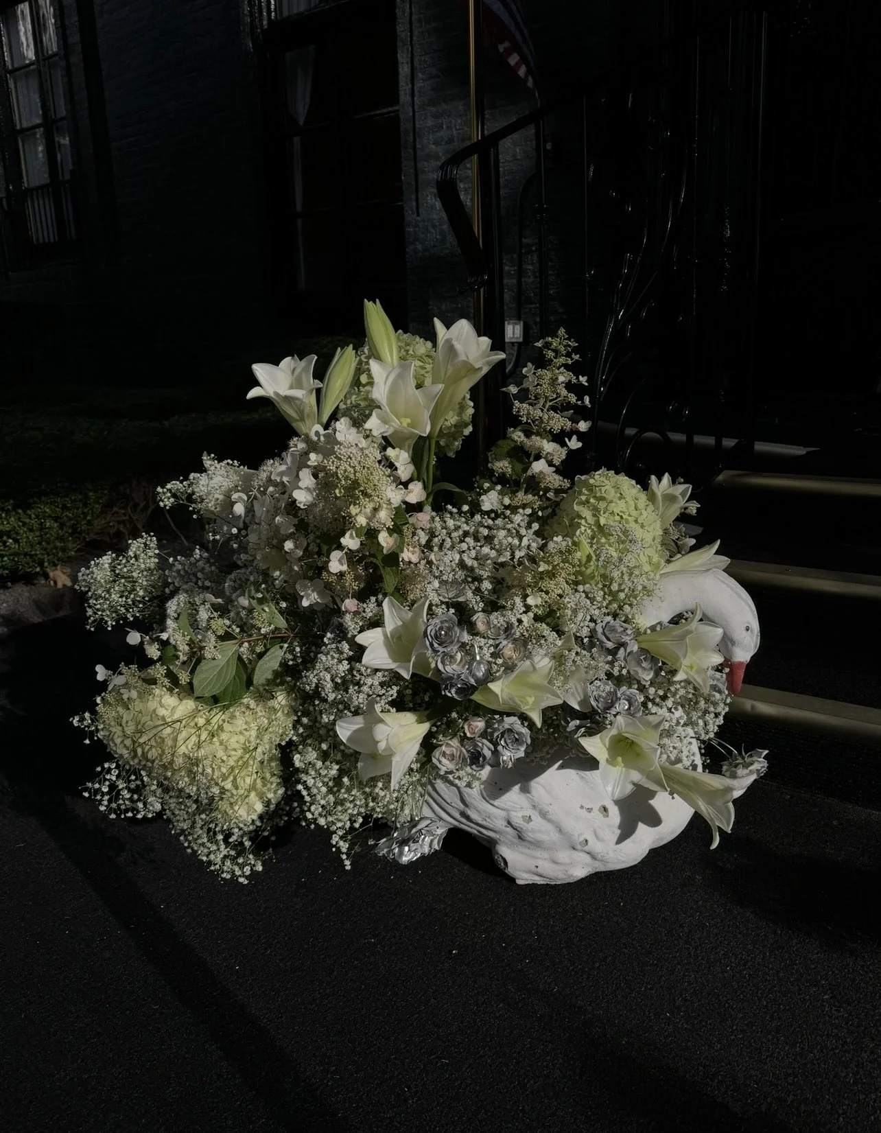 White floral arrangement in a Swan-shaped vase on a dark surface outside at night.