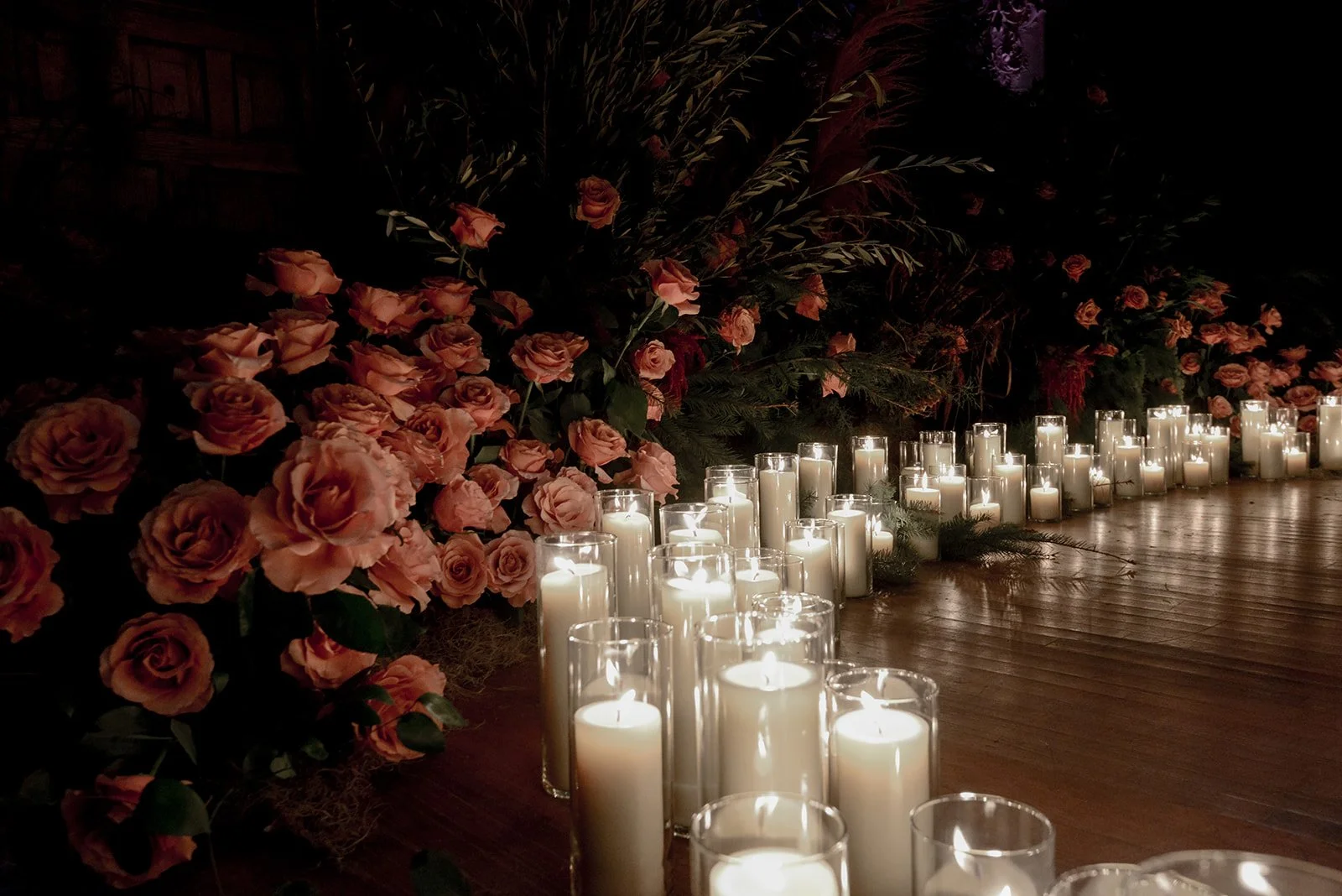 Row of lit white candles in glass holders on wooden floor, surrounded by pink roses and lush greenery for a romantic or memorial setting.