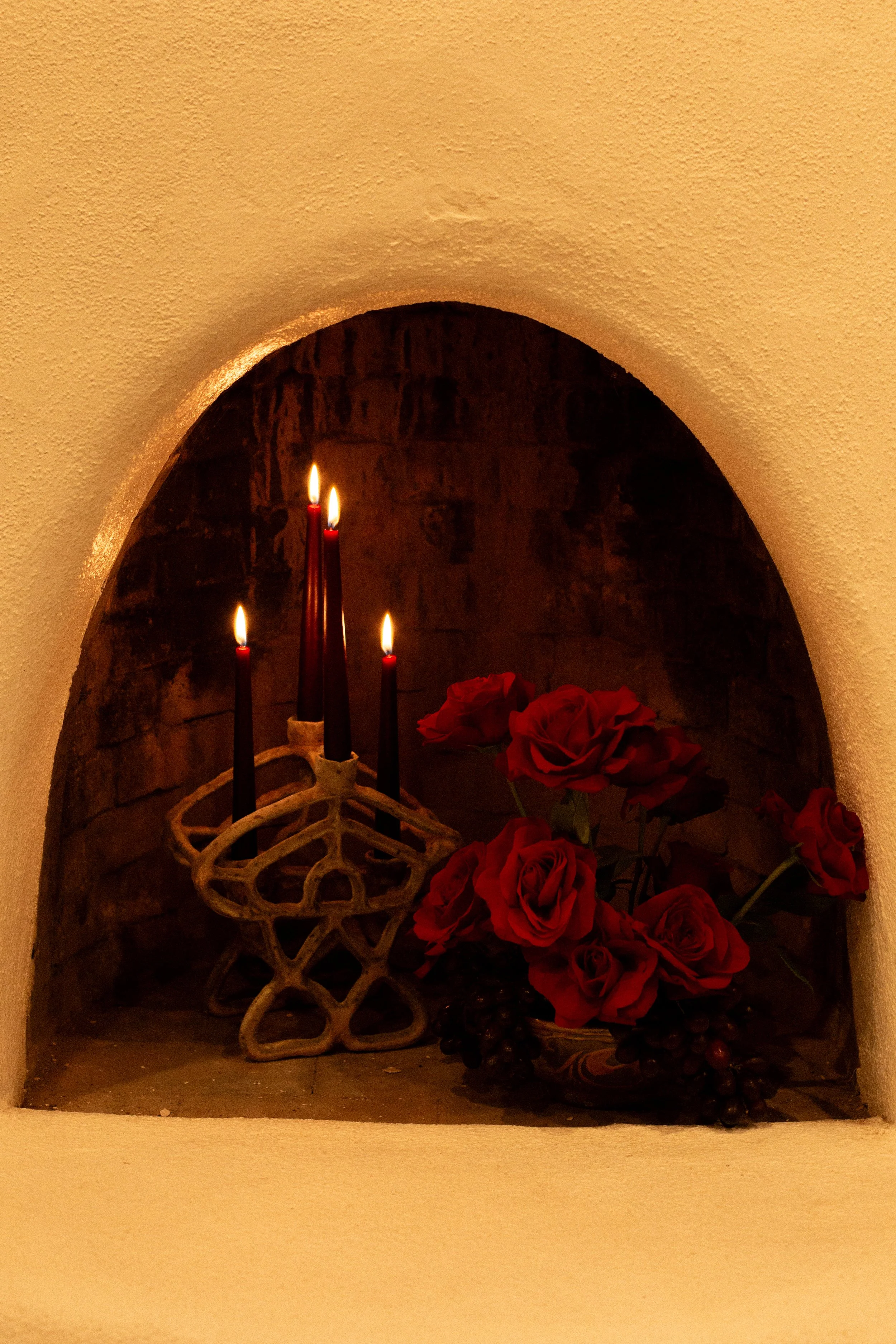 A small alcove with three lit red candles and a bouquet of red roses with some grapes.