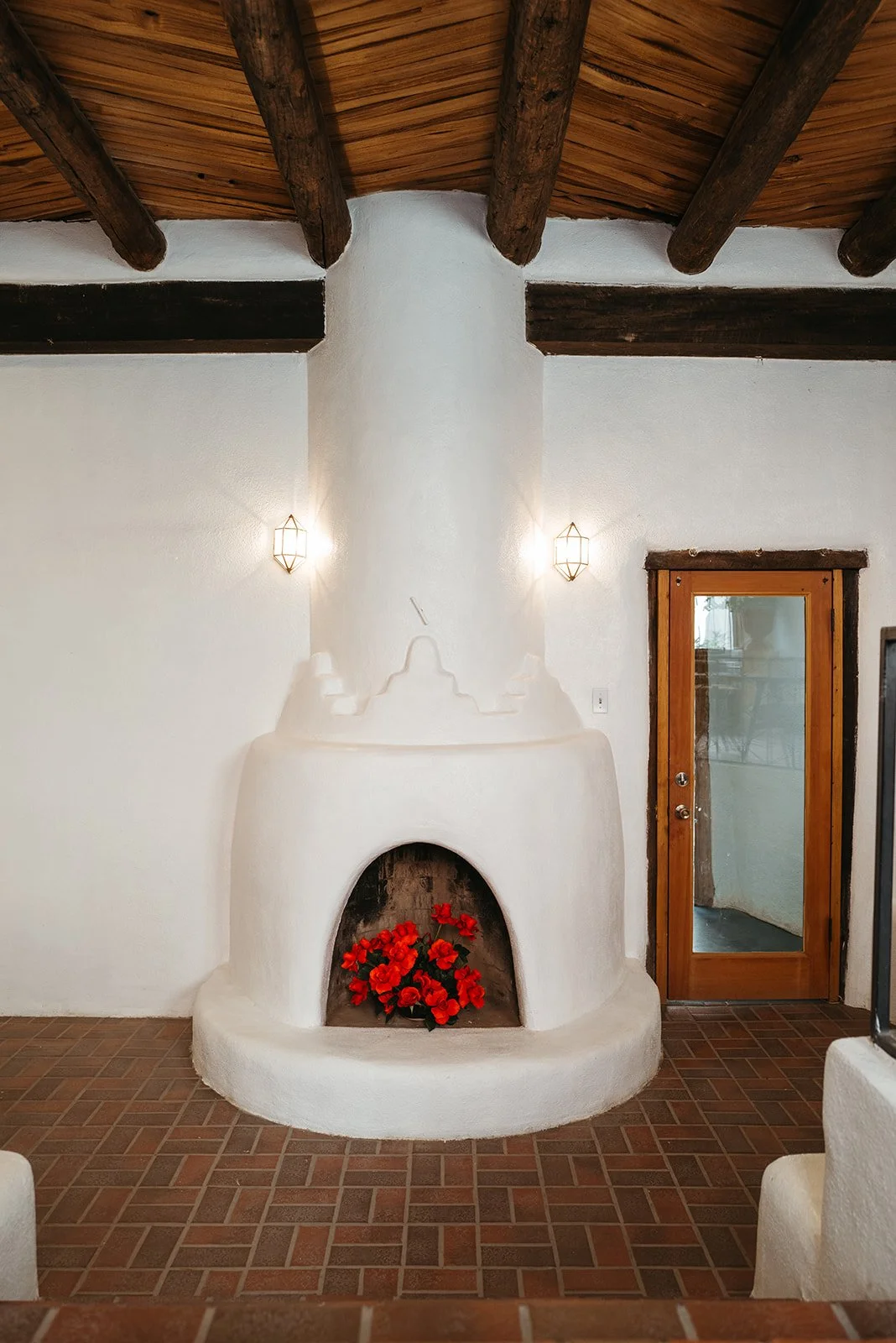 A white stucco fireplace with red flowers inside, rustic wooden ceiling beams, two wall-mounted lights, and a wooden door with glass panel.