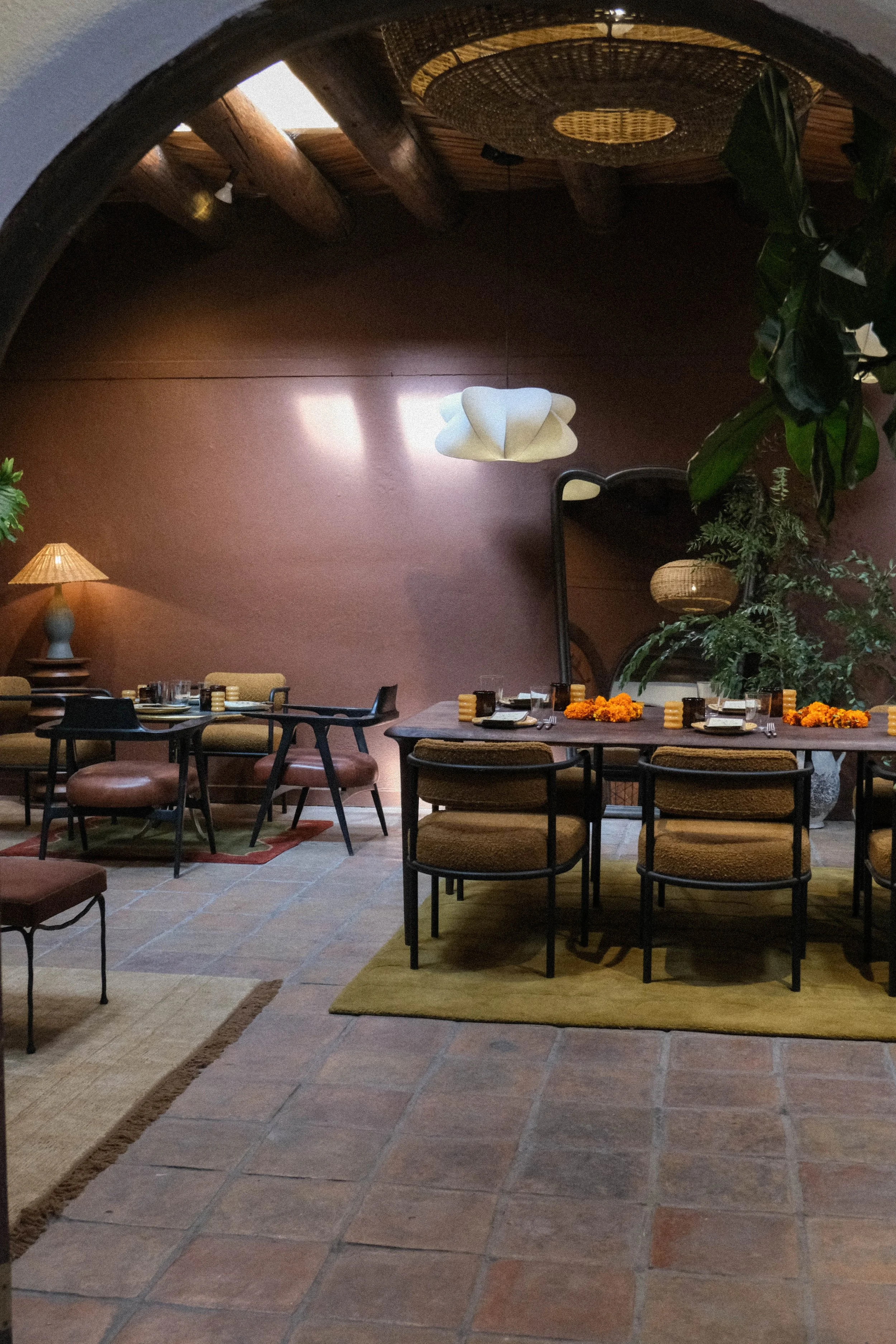 A cozy dining area with a large wooden table decorated with orange marigold flowers, surrounded by cushioned chairs, with a brown wall, a curved mirror, and plants. There is a smaller table with chairs and a lamp, and unique hanging light fixtures.