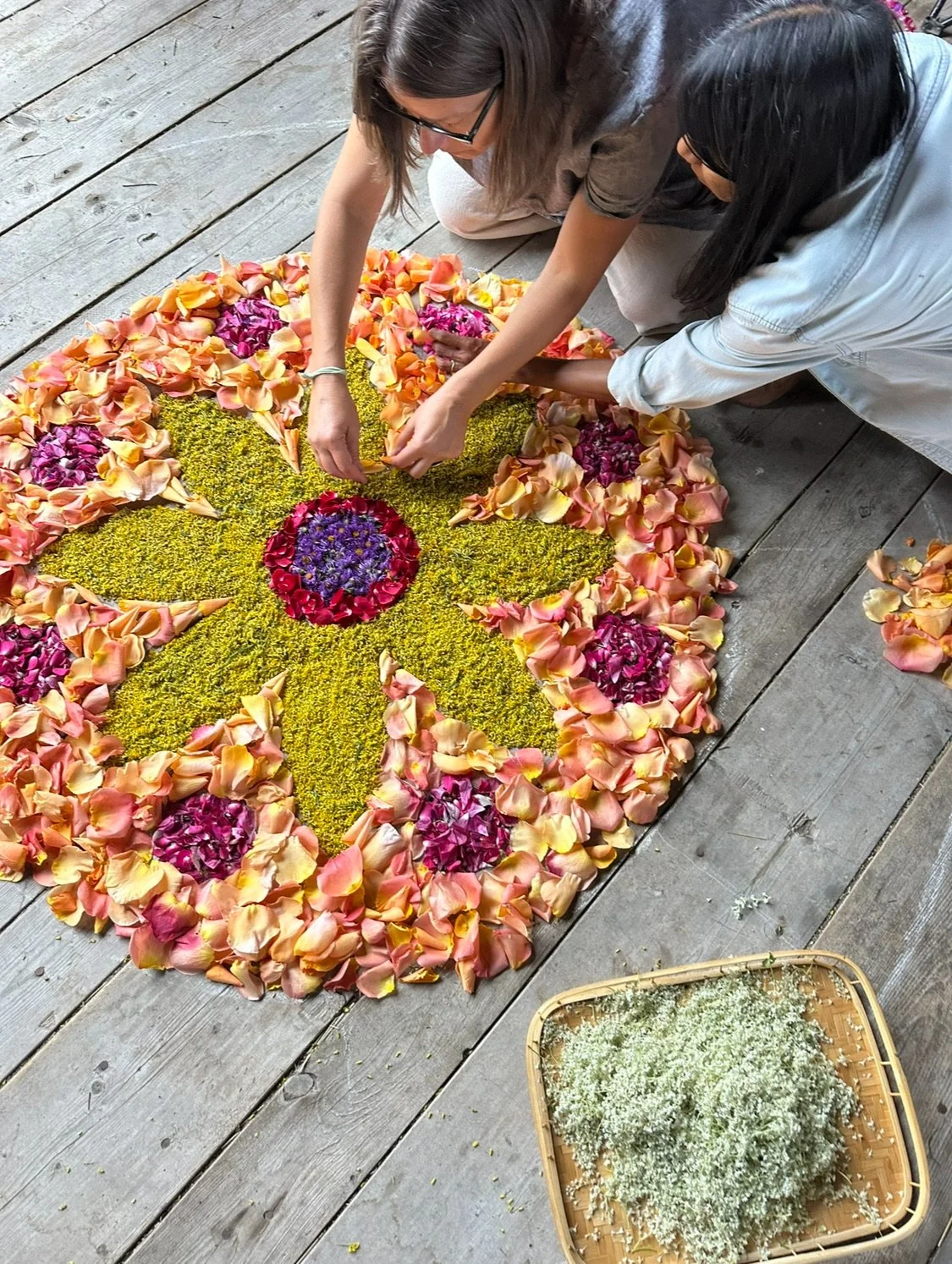 Two women are creating a floral mandala on a wooden floor, using various colorful flowers and petals for decoration.
