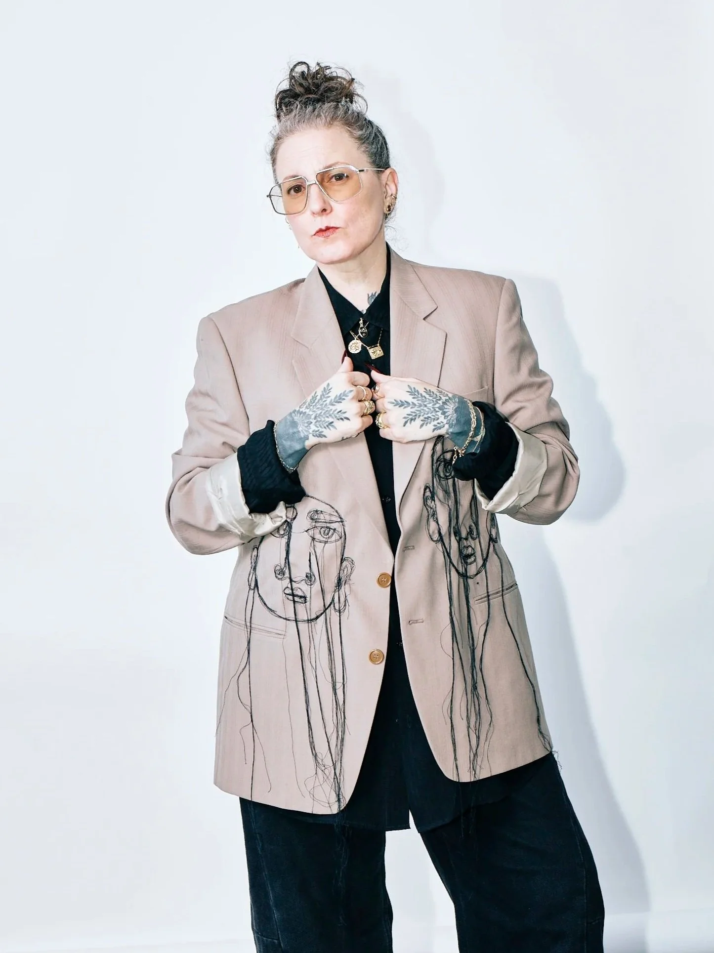 Woman with glasses and tattoos wearing a beige blazer with artistic face line drawings, black shirt, and black pants, posing against a plain white background.