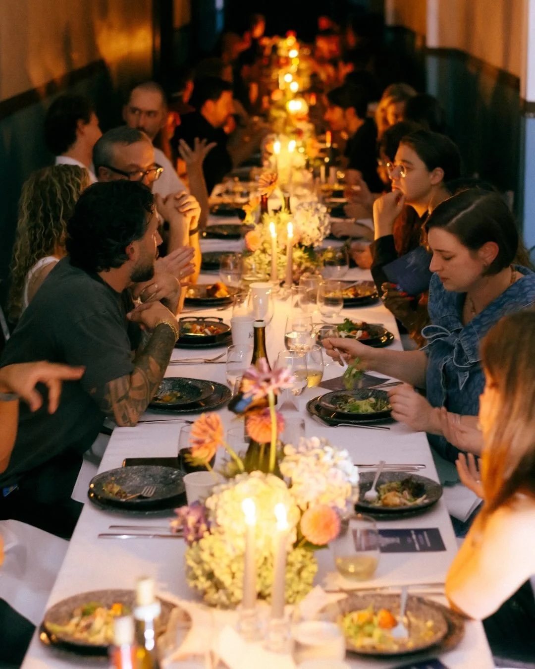 People gathered around a long dining table in a dimly lit restaurant, enjoying a dinner with candles and floral centerpieces.