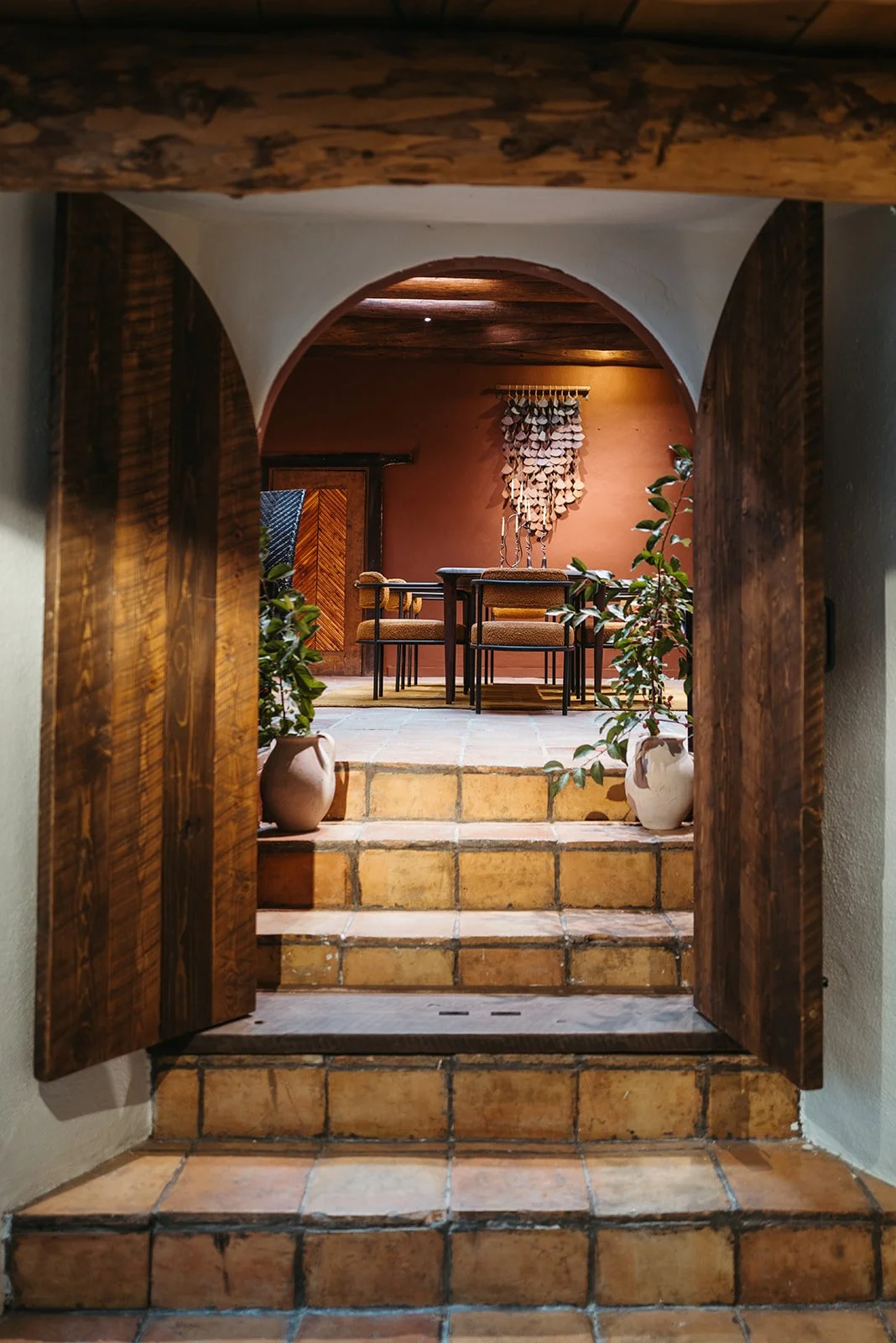 View through an arched doorway into a cozy dining area with a wooden table, chairs, potted plants, and decorative wall art.