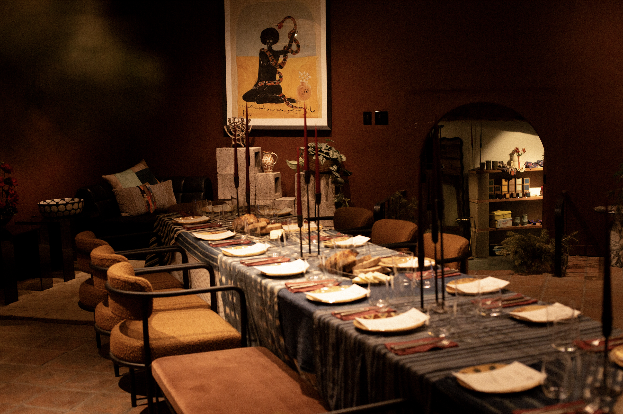 A cozy dining room with a long table set for a meal, surrounded by various chairs, in warm lighting. The wall is painted a deep red, decorated with a framed artwork of a figure with a hoop and a candle. There are shelves with decorative items and pla