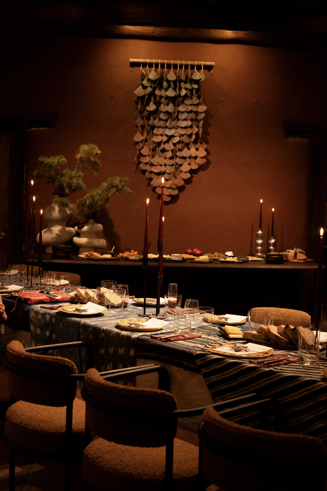 A dimly lit dining room with a long table set for a meal, candles, and a decorative wall hanging made of upside-down clay or cement cups.
