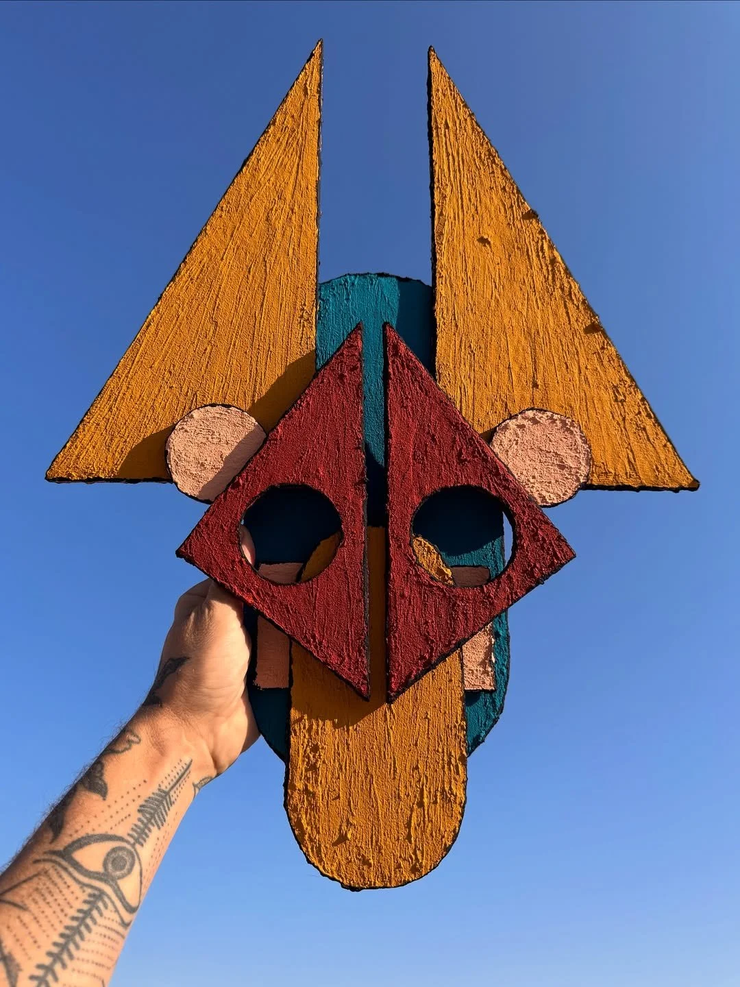 A hand holding a colorful wooden mask with geometric shapes against a clear blue sky.