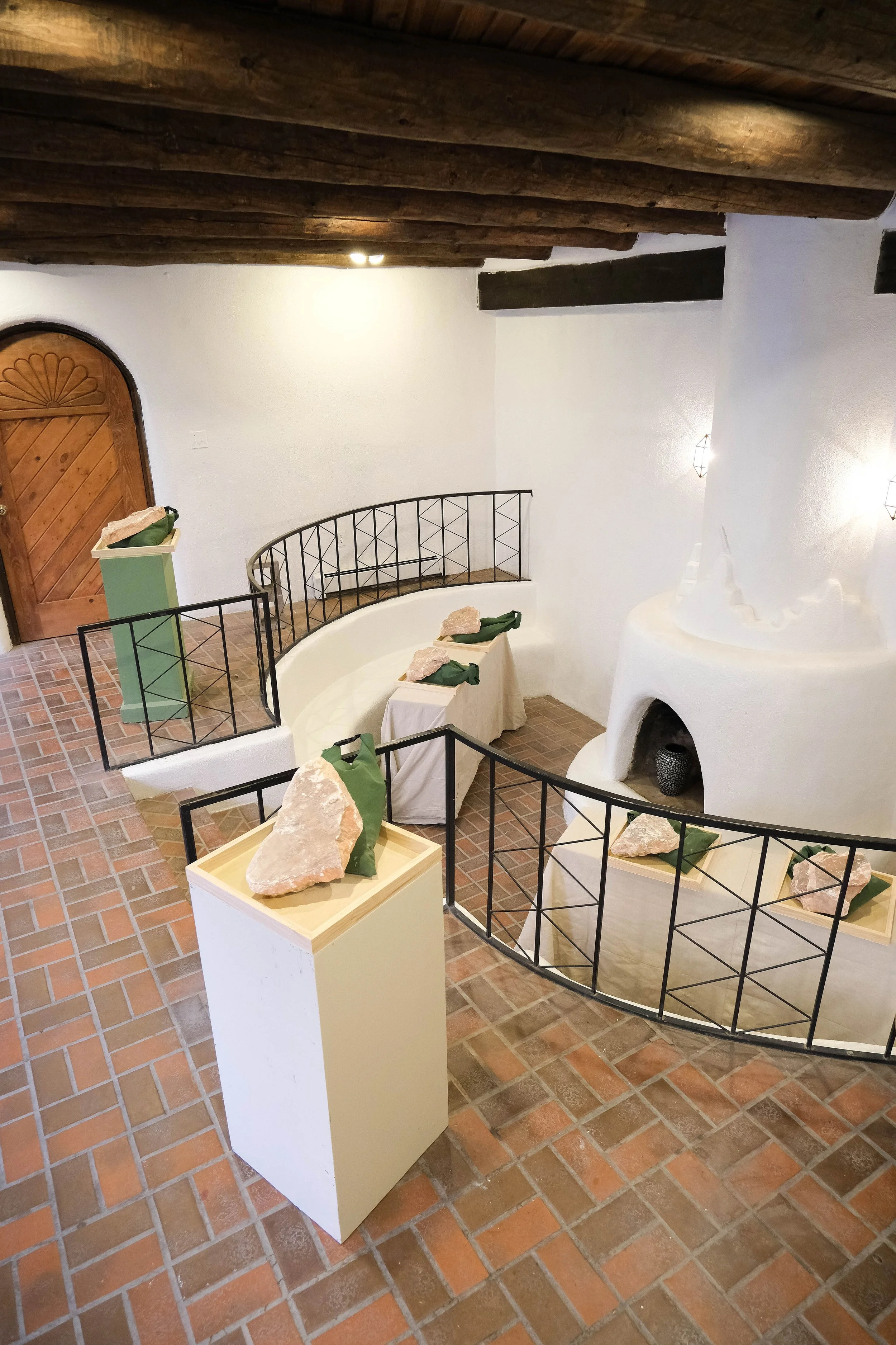 Interior of a decorative room featuring a white, rounded fireplace, black metal handrails, and display tables with large rocks wrapped in green cloth and leaves. The room has brick flooring, white walls, and a wooden ceiling with exposed beams.