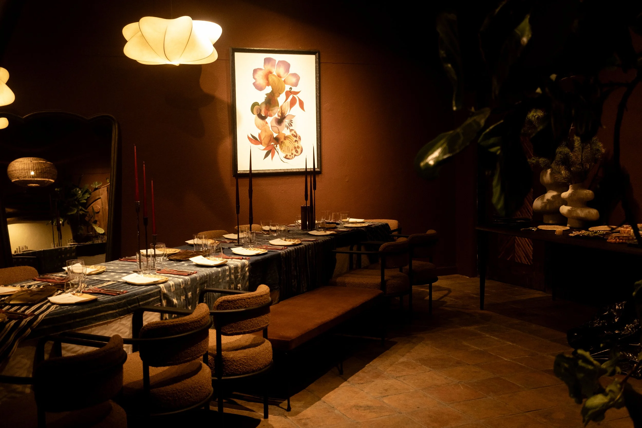 Cozy dining room with a long table set with plates, glasses, and cutlery, surrounded by chairs, and illuminated by warm overhead lighting, featuring a framed floral artwork on the wall.