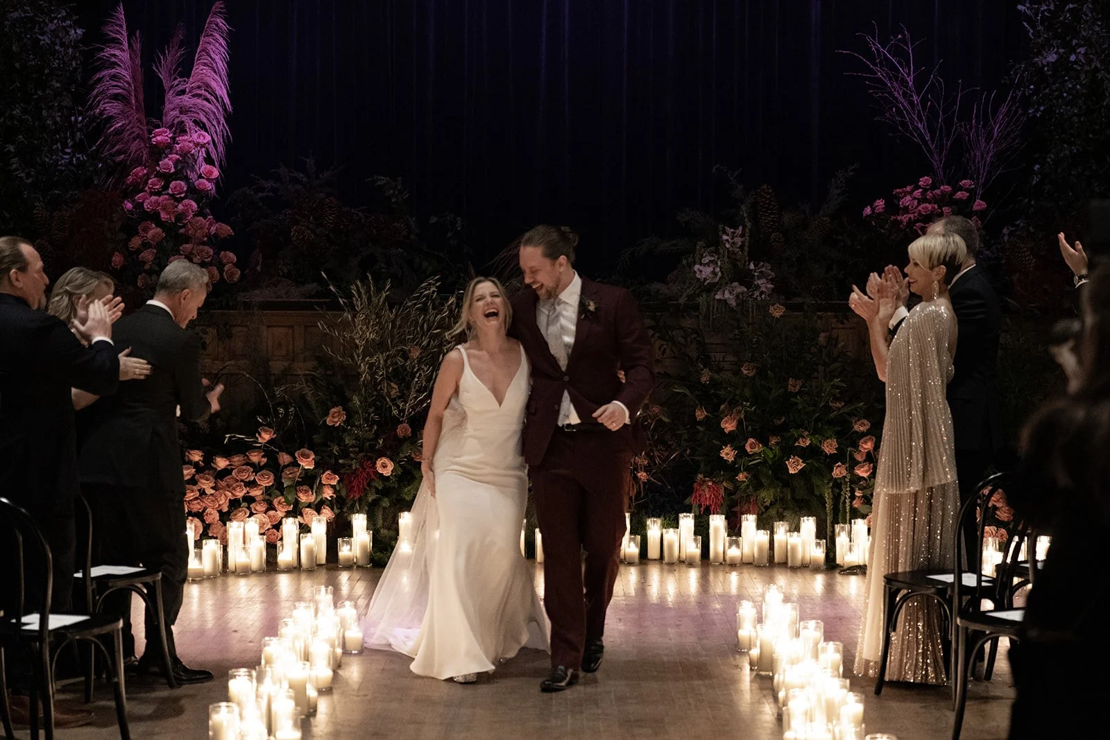A wedding ceremony with a bride and groom walking down the aisle, smiling and laughing, surrounded by candles and floral arrangements, with guests clapping and celebrating.