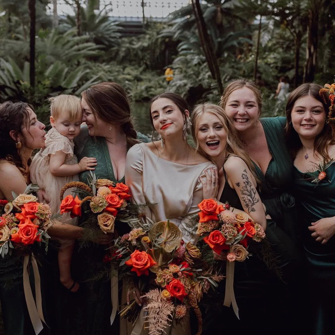 A group of women and children smiling together, holding bouquets of orange and cream flowers, in a lush indoor greenhouse.