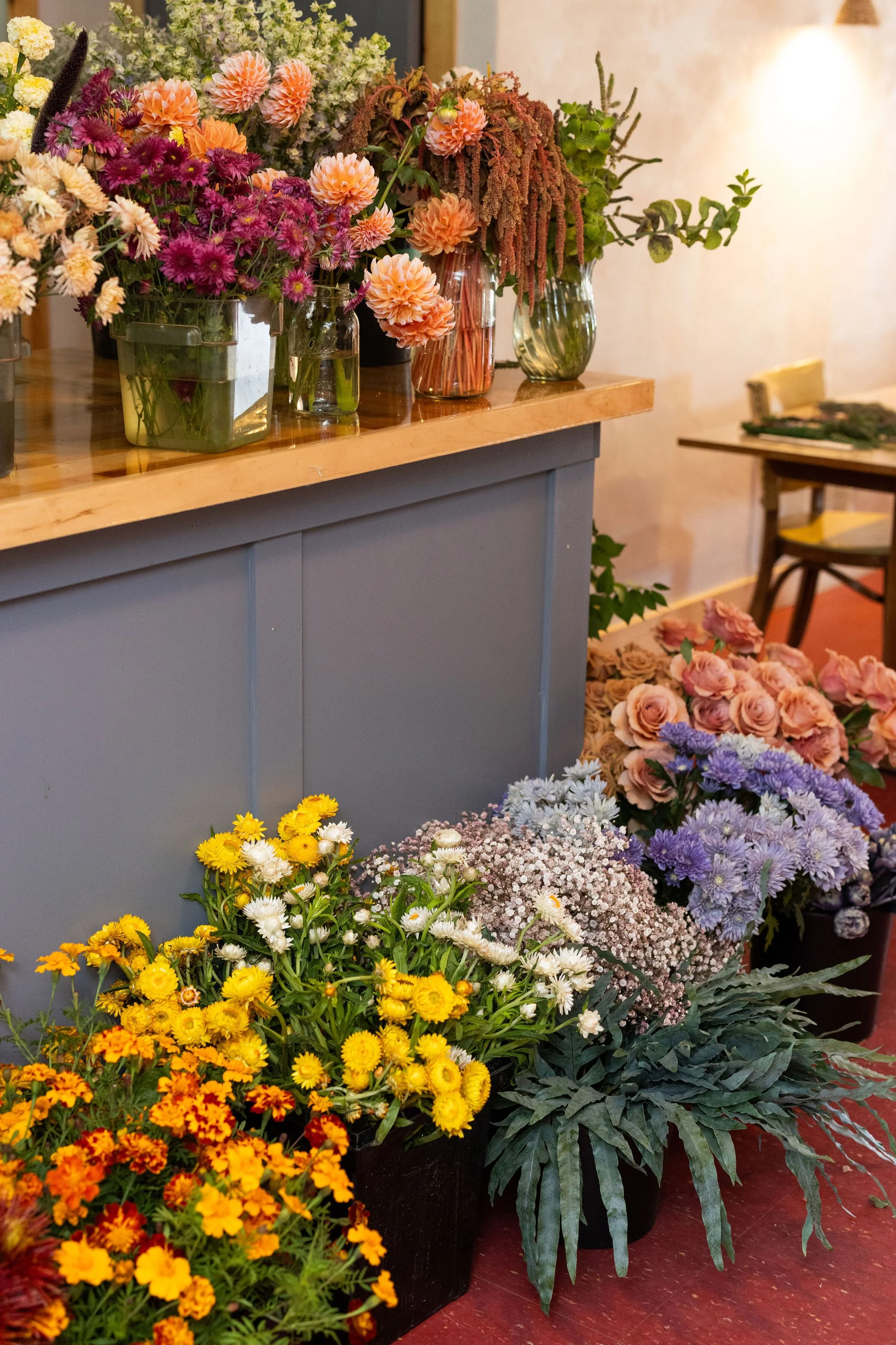Various colorful flowers in vases and pots, including mums, roses, and other blooms, arranged on a wooden table and on the floor.