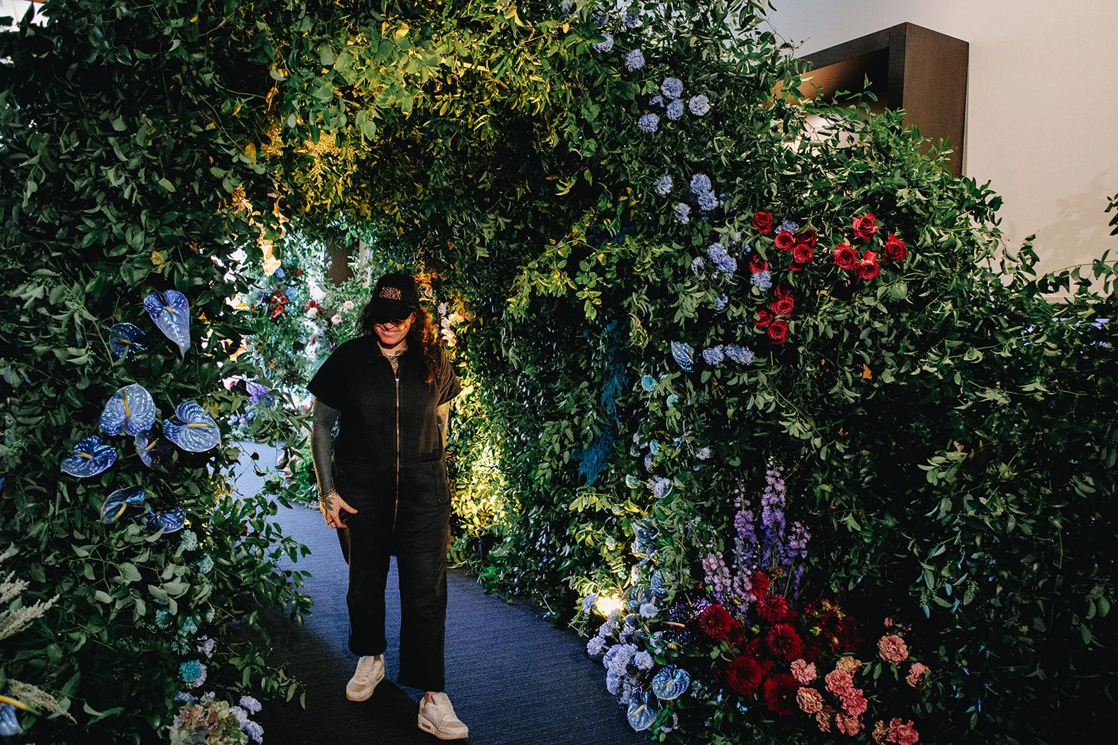 A woman walking through a floral archway decorated with various colorful flowers and lush green foliage, with moon-style lighting highlighting the scene.