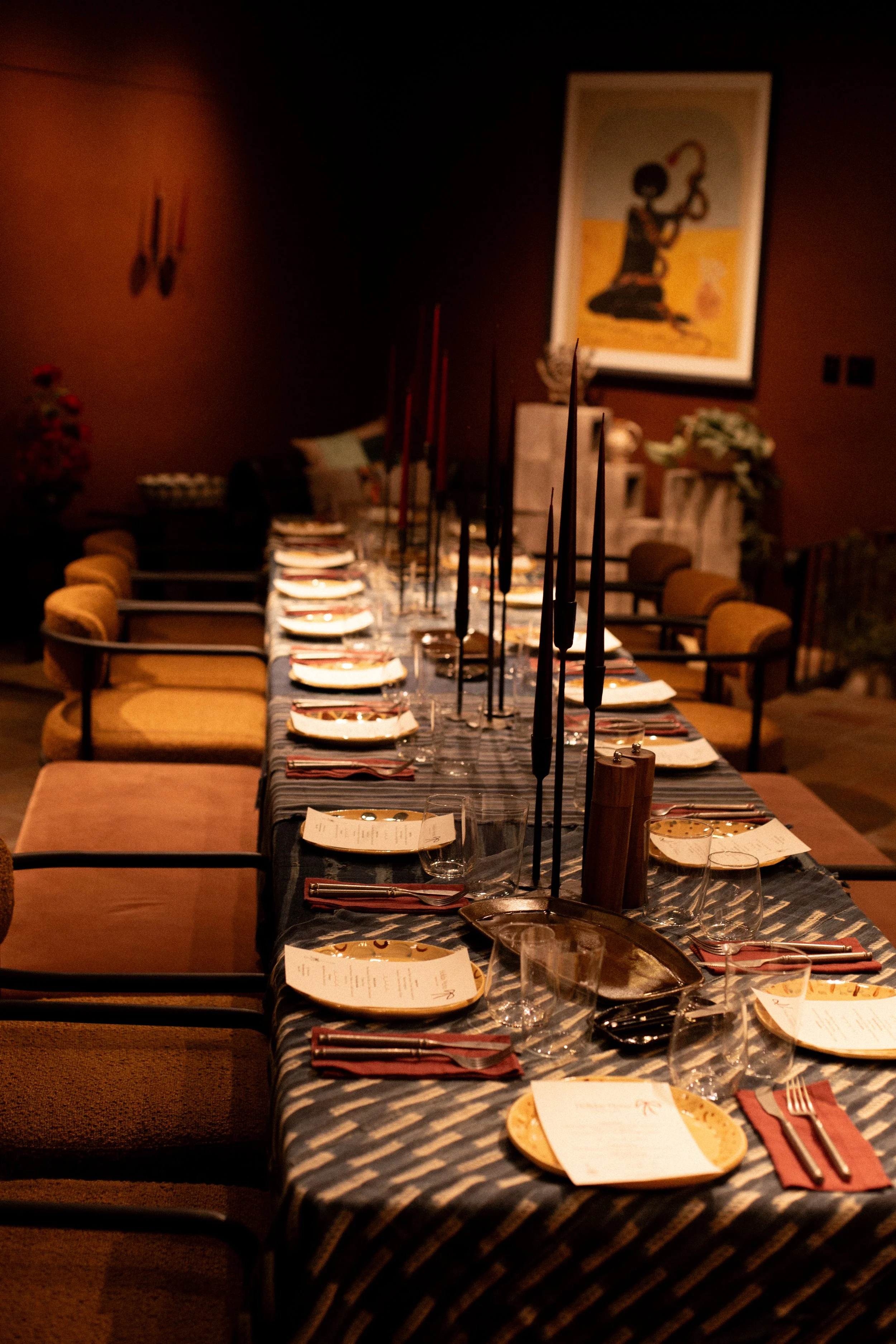 A long dining table set for a meal with plates, glasses, silverware, and elongated black candles. The table has a dark cloth and is in a dimly lit room with wall art and chairs around.