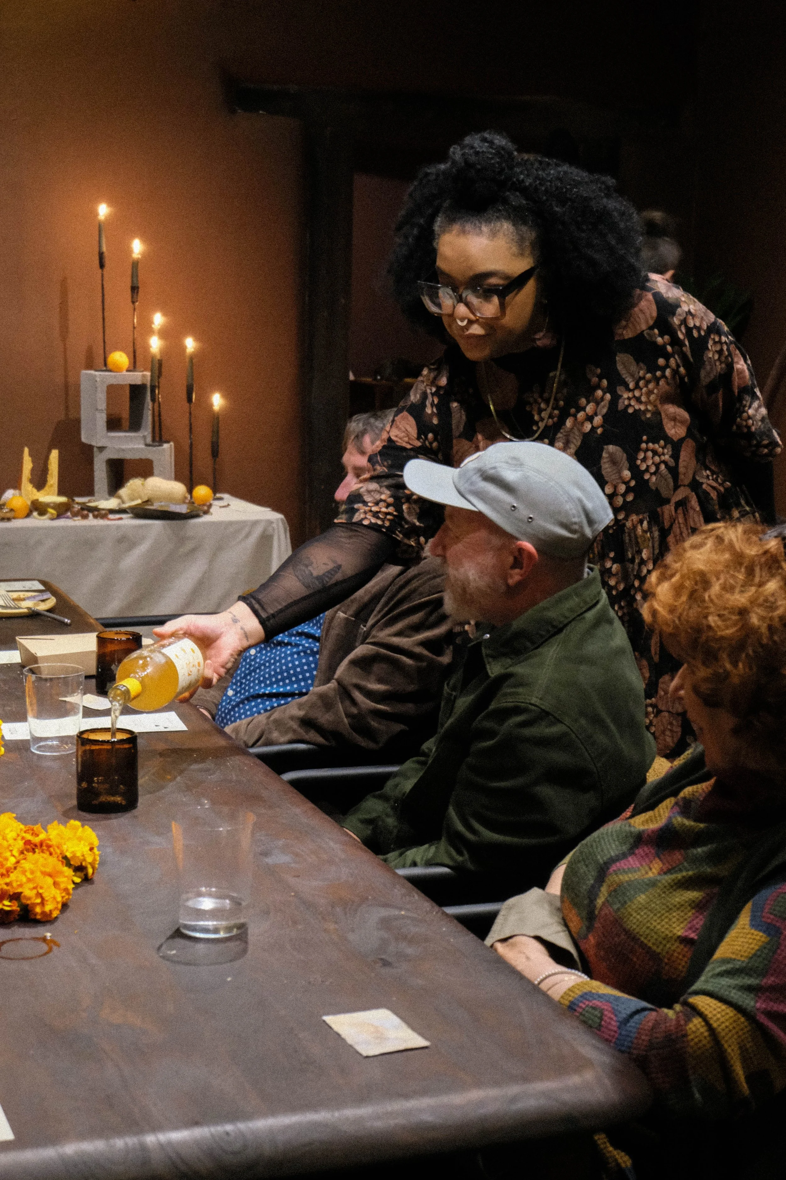 A woman with glasses and black curly hair pours a yellow beverage into a glass at a dinner party. Other guests are seated at the table, and there is a decorated table with candles and yellow flowers in the background.