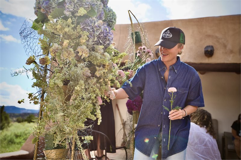 A person in a blue shirt and baseball cap smiling and holding a pink flower, standing near large floral arrangements outdoors.