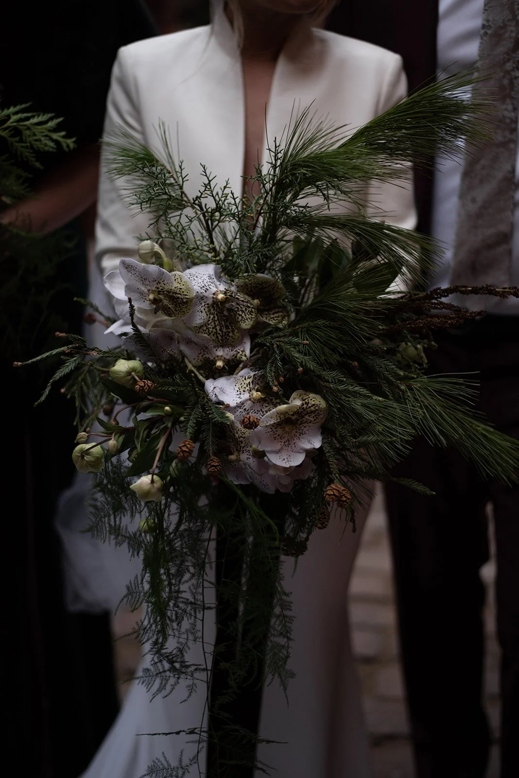 A woman in a white blazer holding a large bouquet of greenery and white orchids.