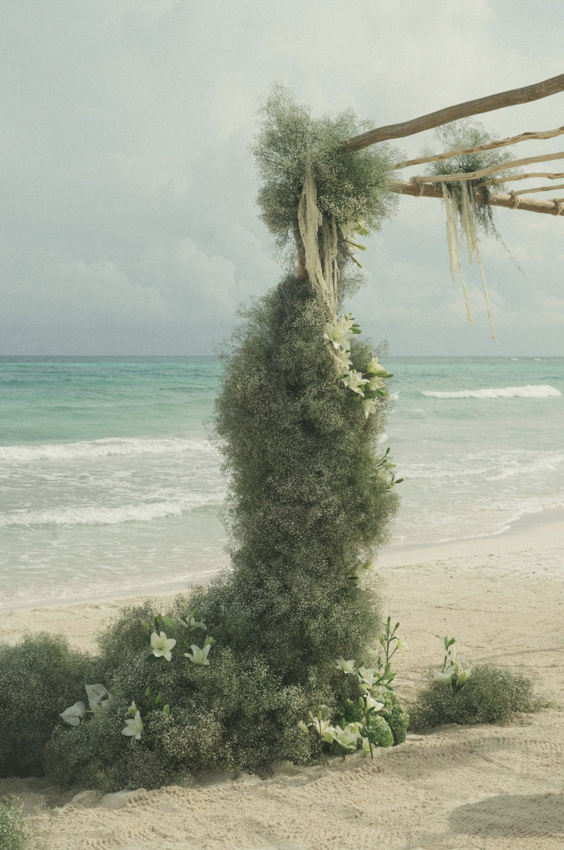 Decorative floral arrangement with greenery and white flowers on a sandy beach, with the ocean and cloudy sky in the background.