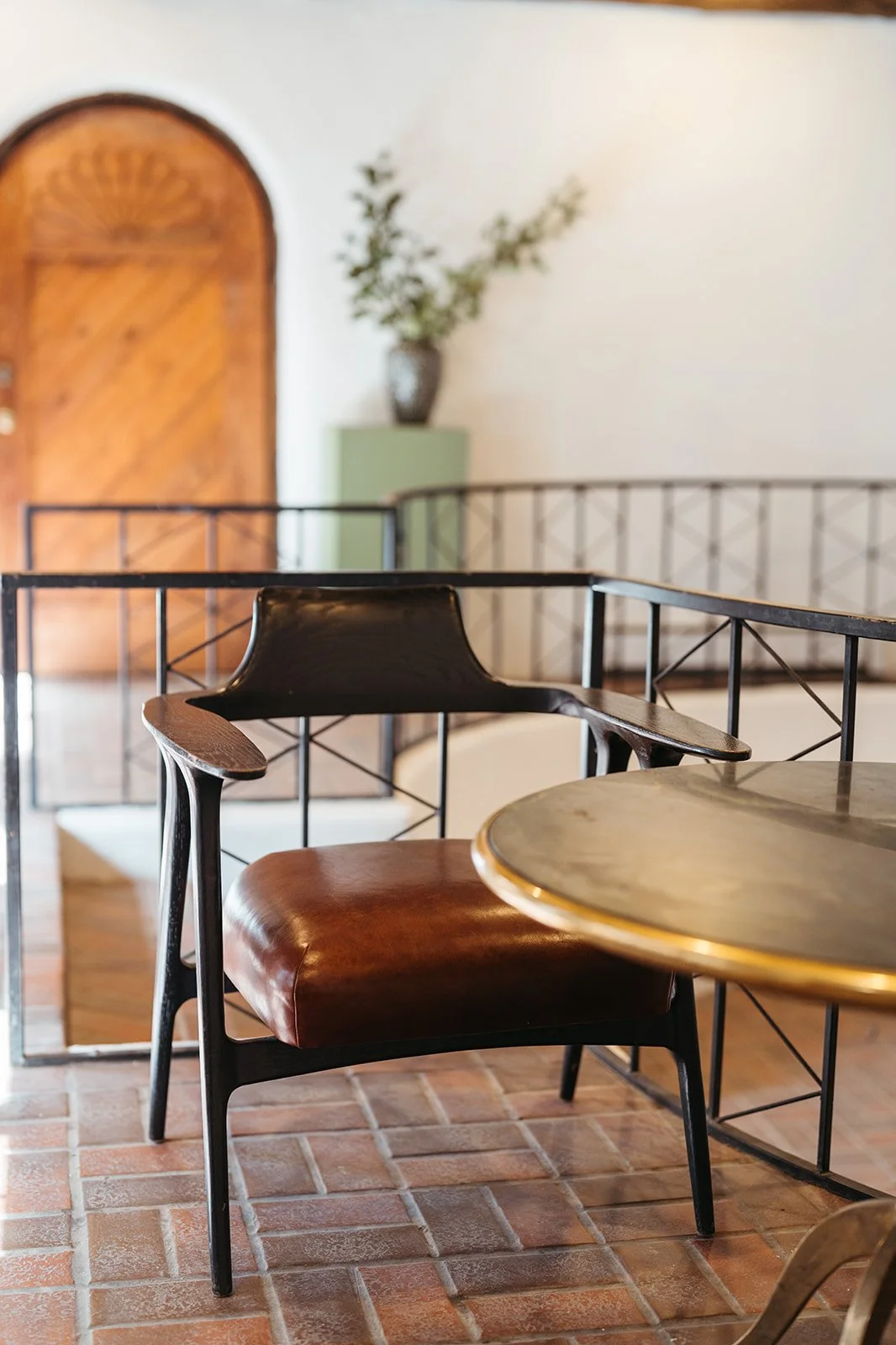 A black and brown wooden chair with a leather seat next to a round table with a brass edge, on a brick floor inside a room with a wooden door, potted plant, and decorative railing in the background.