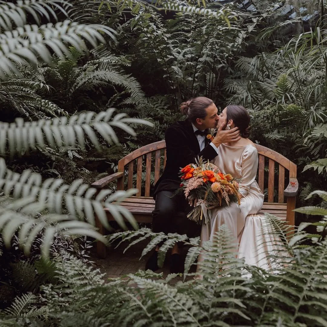 A couple dressed in wedding attire sitting on a wooden bench in a lush, green garden, sharing a kiss. The woman holds a bouquet of orange and beige flowers.