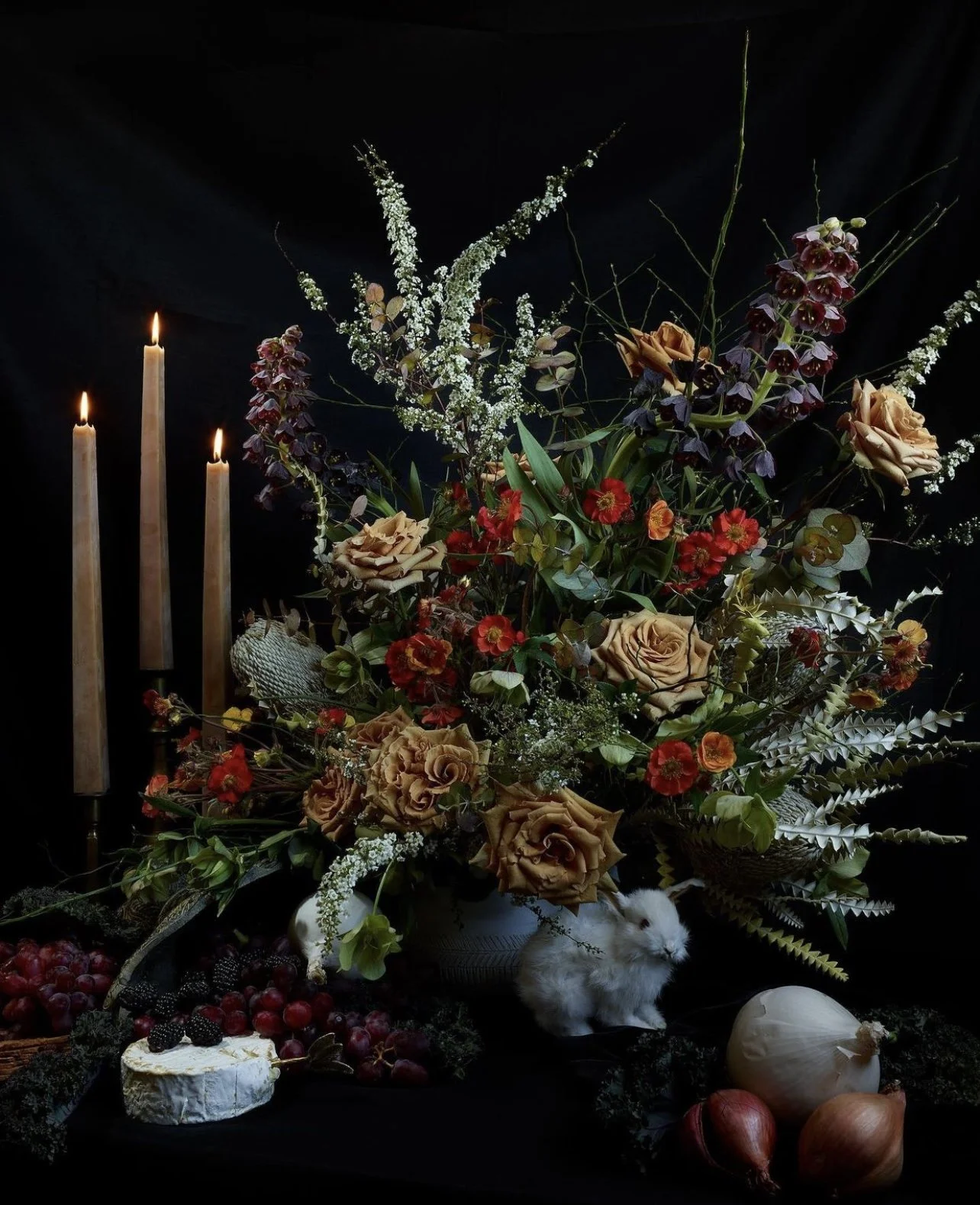 A floral display with beige roses, red and purple flowers, surrounded by greenery, and three lit candles on the left, all set against a black background, with fruit and a small white rabbit at the bottom.
