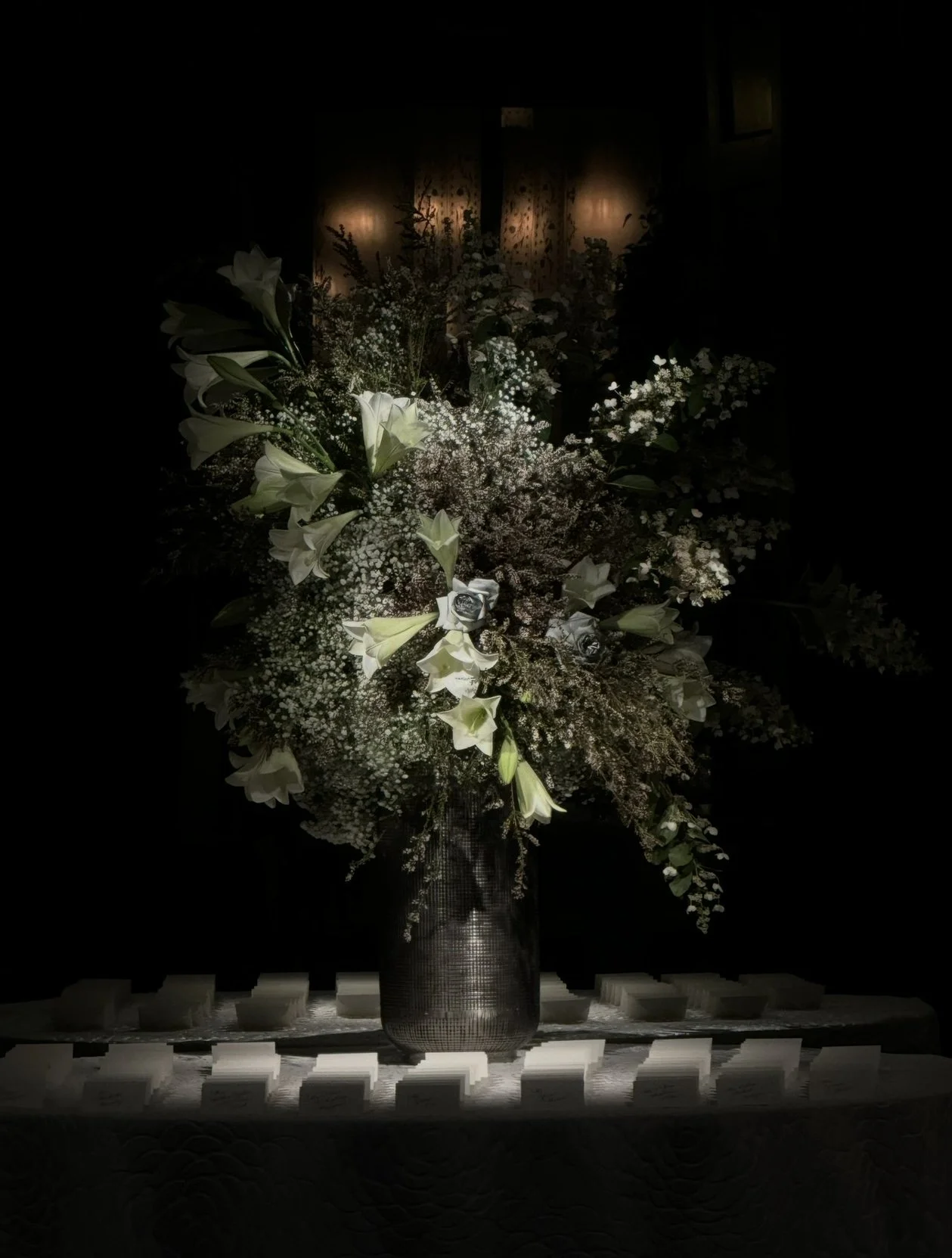 A dark room with a large bouquet of white flowers, including lilies and baby's breath, illuminated by soft lighting, placed in a metallic vase on a table with white cards or booklets arranged in front.