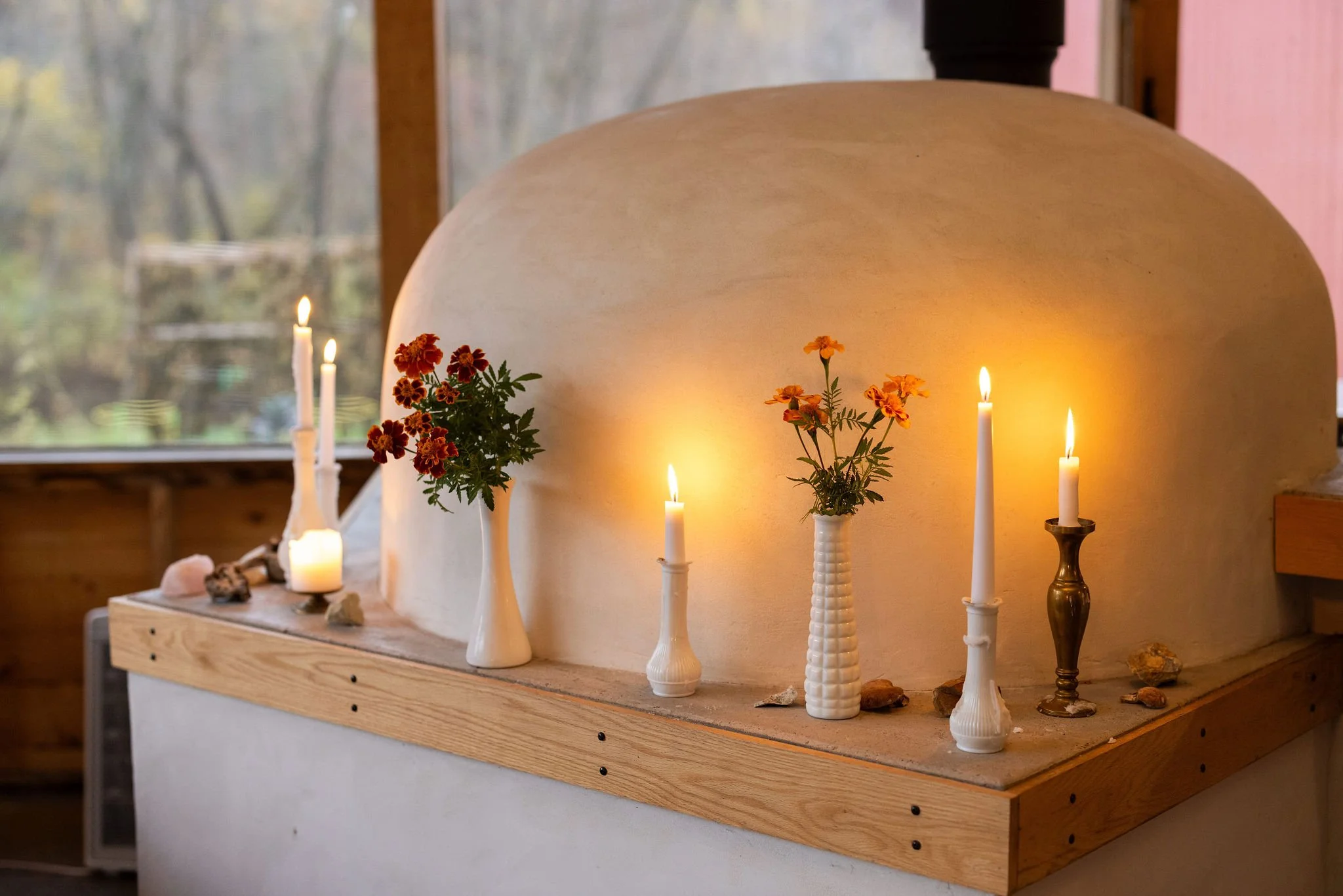 Candles in vases and a brass candle holder with flowers on a wooden shelf, with a large rounded structure and a window in the background.