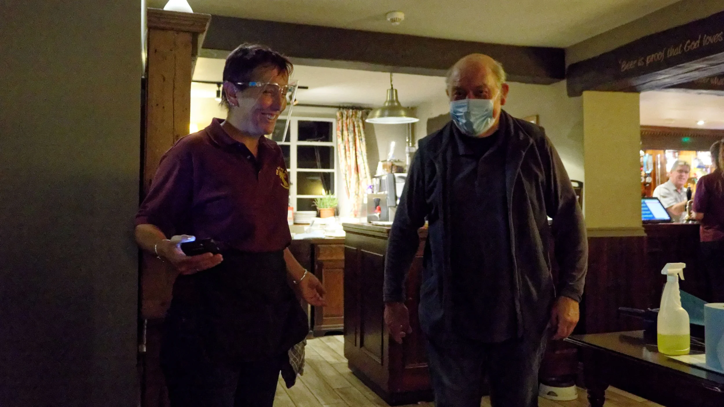 Woman guides customer to his seat in the pub. Both wear masks.