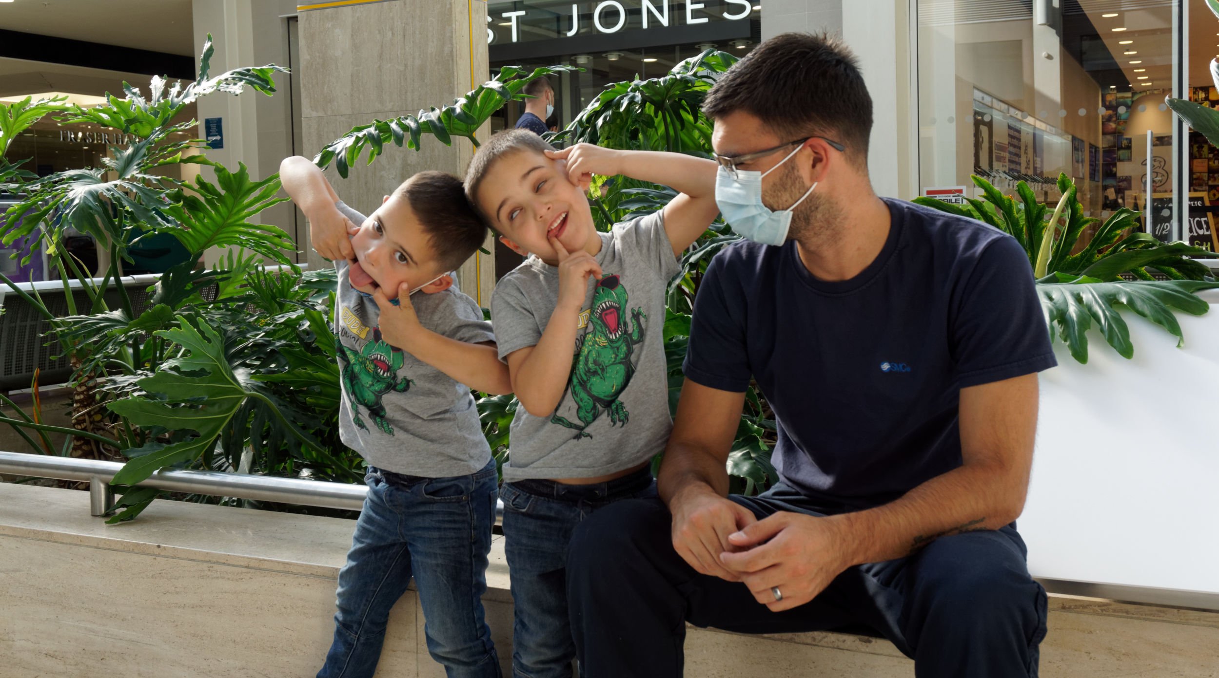 Twin boys remove their Covid masks to pull faces; father's amused.