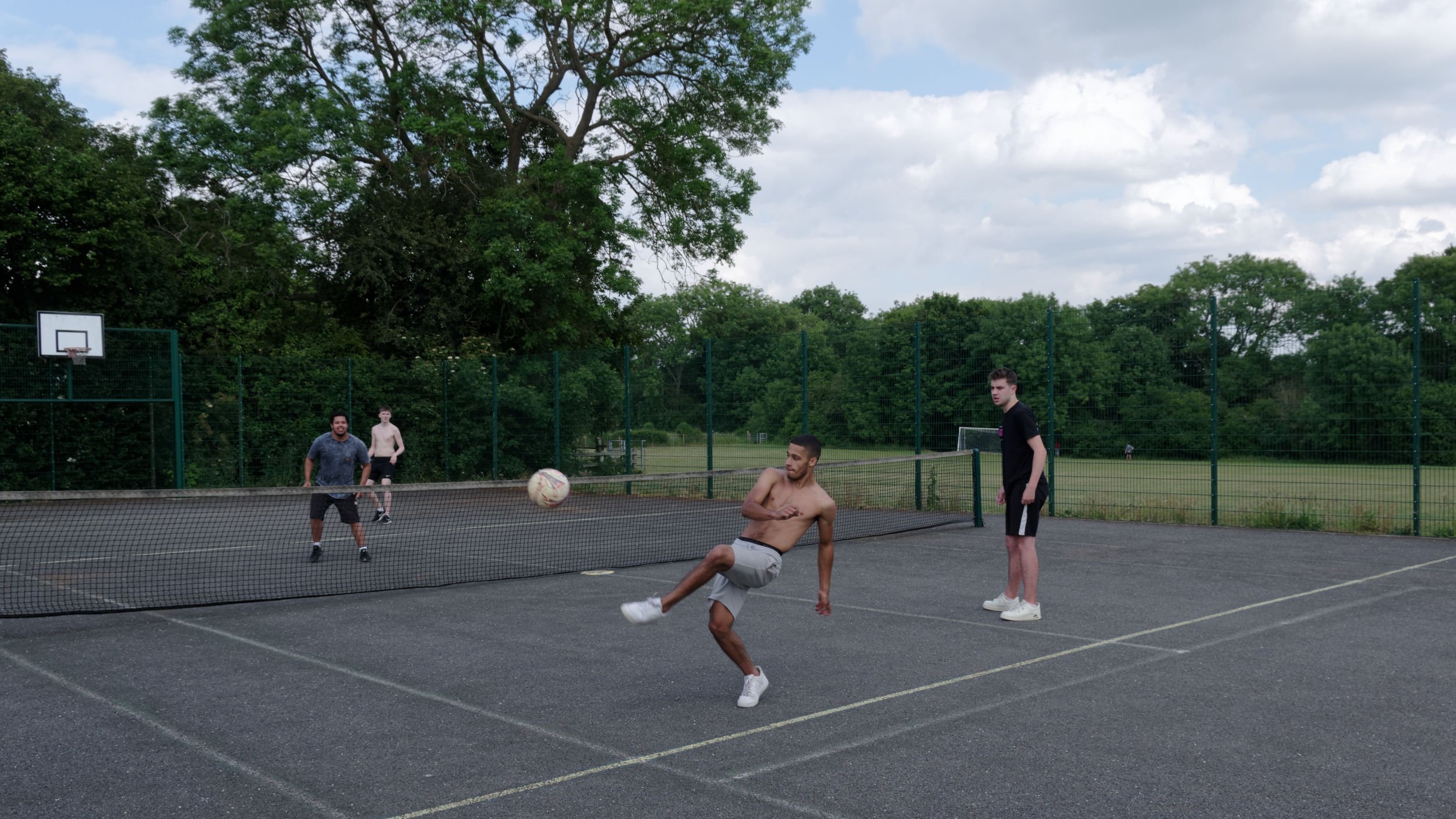 Social distancing youths kick a football around a tennis court.