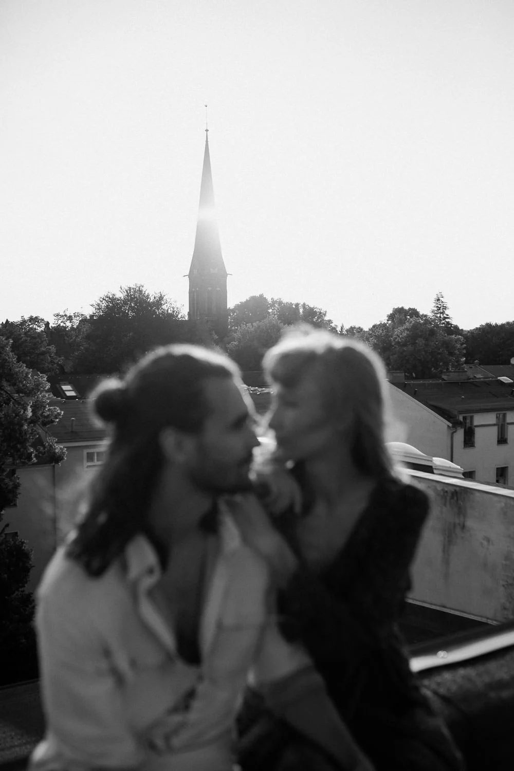 Zwei Menschen, die auf einem Balkon nahe beieinander sitzen und sich küssen, mit einer Kirche und Bäumen im Hintergrund, im Schwarz-Weiß-Stil.