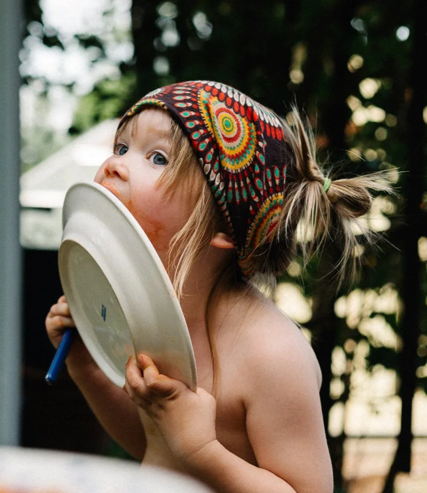 Ein Kind mit buntem Kopftuch leckt einen Teller ab.