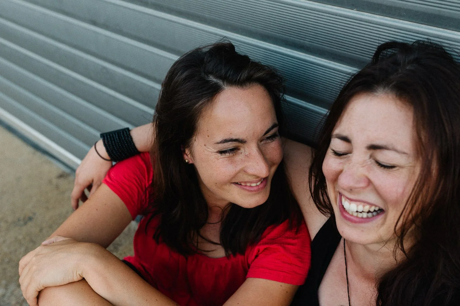 Zwei lachende Frauen sitzen an einer Metallwand, eine trägt ein rotes T-Shirt, die andere ein schwarzes Top, und sie genießen einen Moment der Freude und Freundschaft.